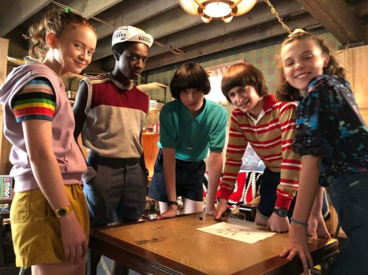 Stranger Things Funny 桌布 壁紙 — 五個孩子靠在一張木桌上，在一個質樸的閣樓裡，微笑著專注於地圖或圖畫。