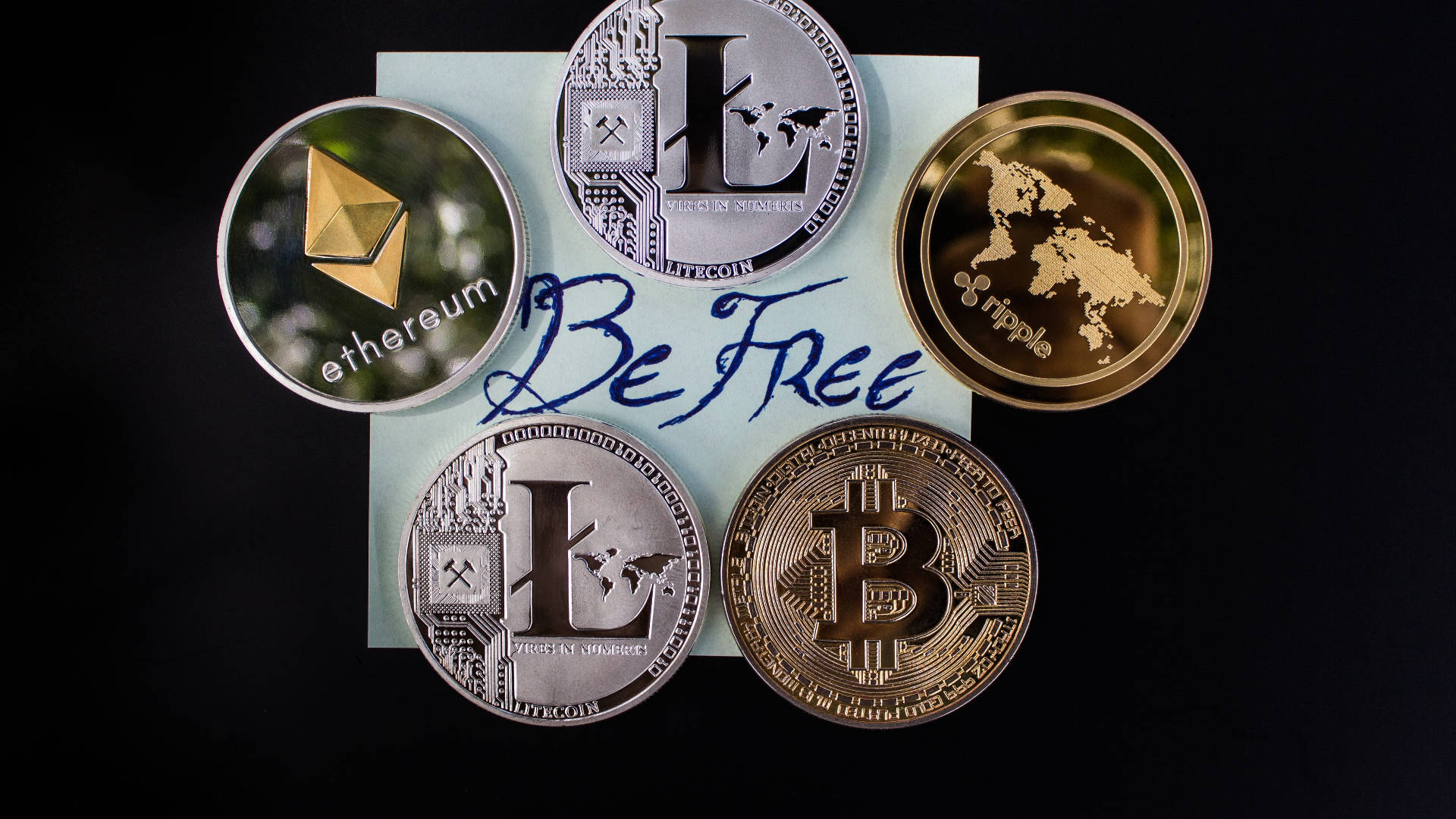 以太坊 桌布 壁紙 — 五個實體加密貨幣硬幣（Ethereum、Litecoin、Bitcoin 和 Ripple）圍繞著一張手寫的「Be Free」便條，放在淺藍色紙上，黑色背景。