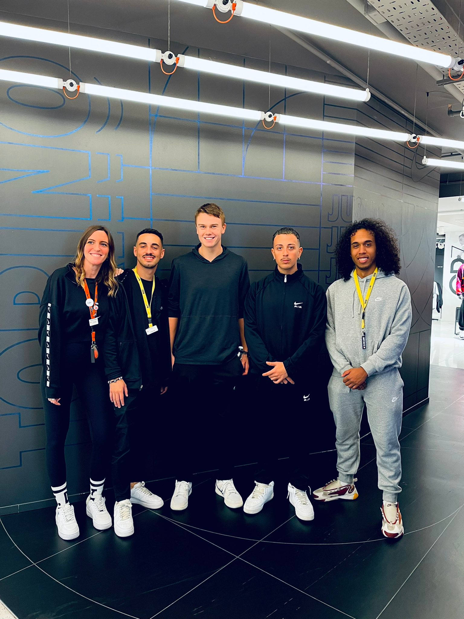 Teamwork 桌布 壁紙 — 五個微笑的人穿著運動服在現代零售店內一起擺姿勢，店內有幾何牆壁藝術和明亮的頭頂燈光。