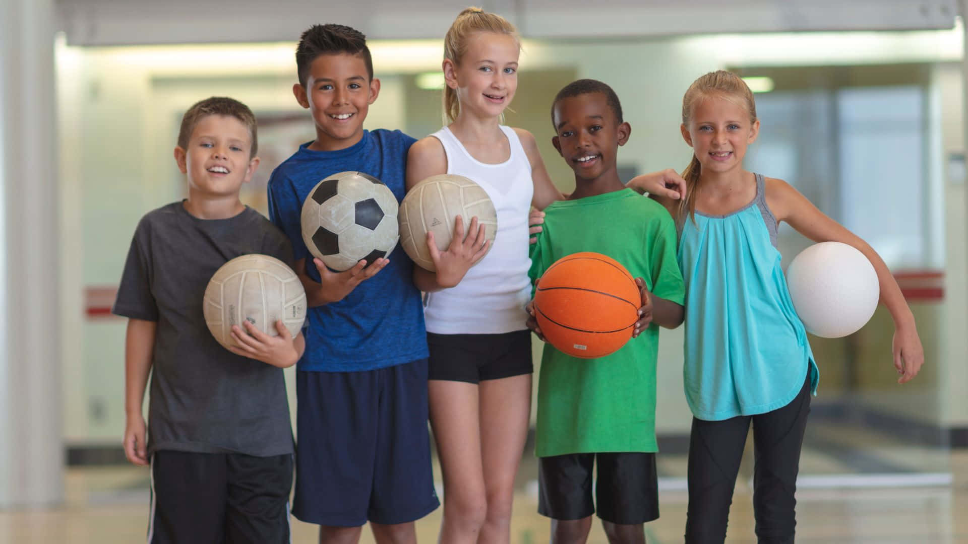 Best Sports 桌布 壁紙 — 五個微笑的孩子穿著運動服，手持各種運動球，包括足球、排球、籃球和一個白球，一起在室內擺姿勢。