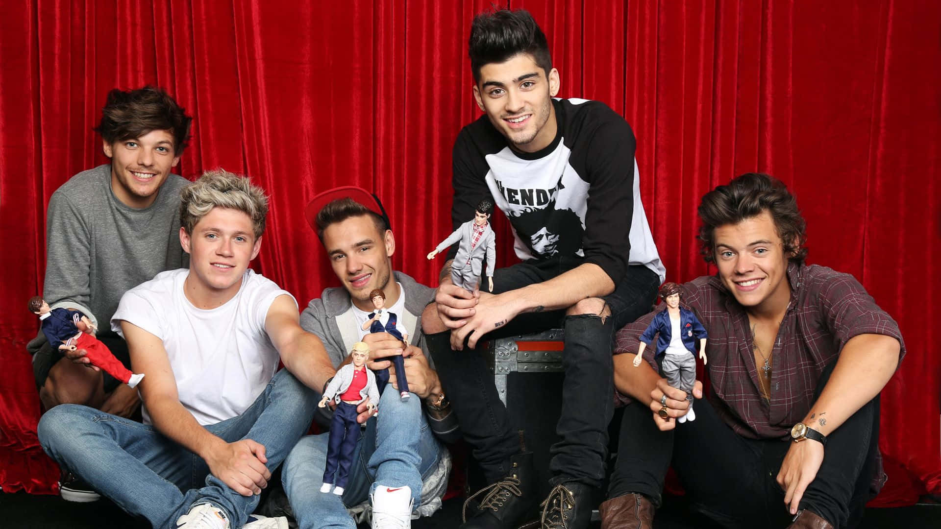 One Direction 桌布 壁紙 — 五個微笑的年輕男子在紅色天鵝絨簾幕的舞台上隨意擺姿勢，手持自己的小型玩偶版本。