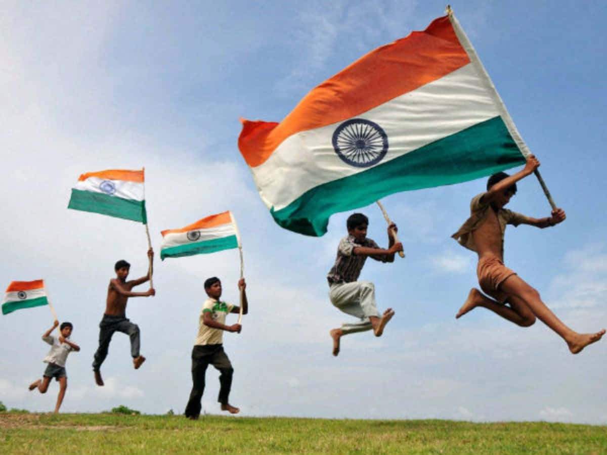 India Independence Day 桌布 壁紙 — 五個快樂的孩子在草地上高高跳起，揮舞著印度國旗，背景是明亮的藍天。
