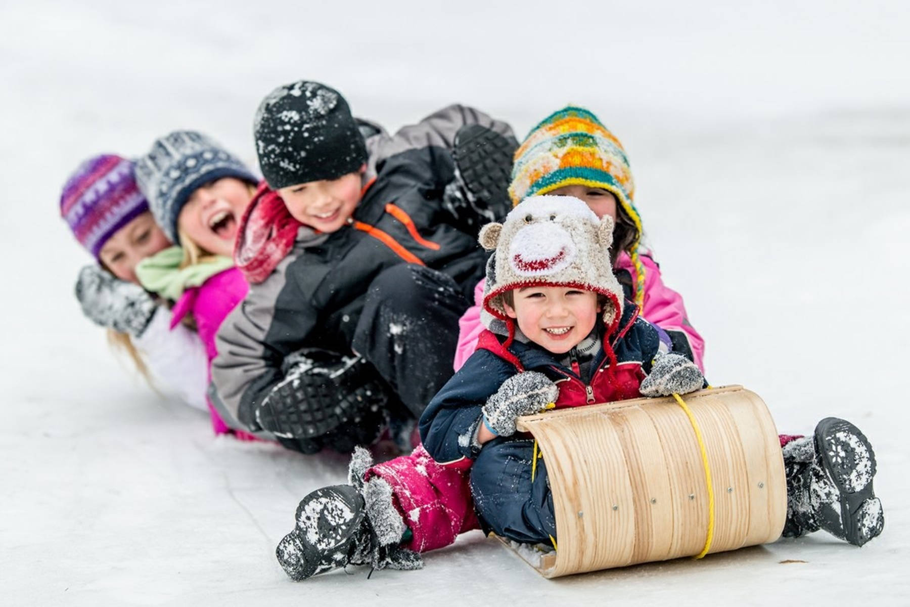 雪 桌布 壁紙 — 五個快樂的孩子穿著色彩繽紛的冬季裝備，在雪坡上滑雪橇，笑聲燦爛。