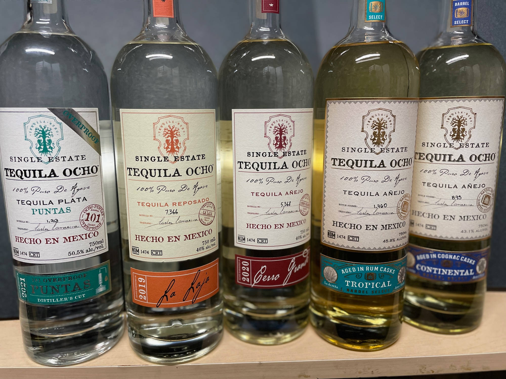 bottle 桌布 壁紙 — 五個標有標籤的龍舌蘭酒瓶，年齡和風格各異，整齊排列在木架上，帶有透明玻璃和彩色標籤。
