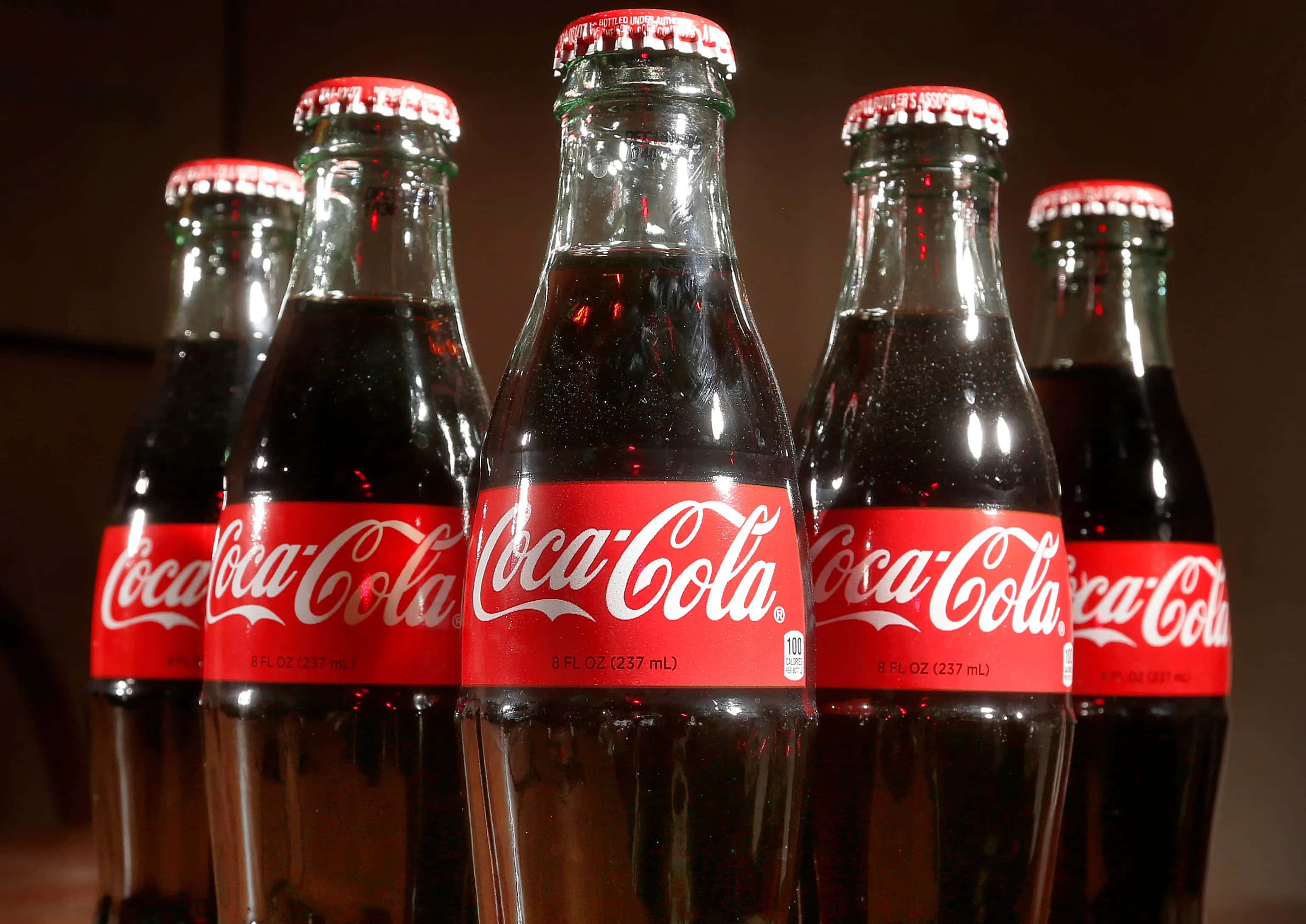 coca cola 桌布 壁紙 — 五個玻璃可口可樂瓶，帶有紅色標籤和瓶蓋，在深色背景上排成一排。