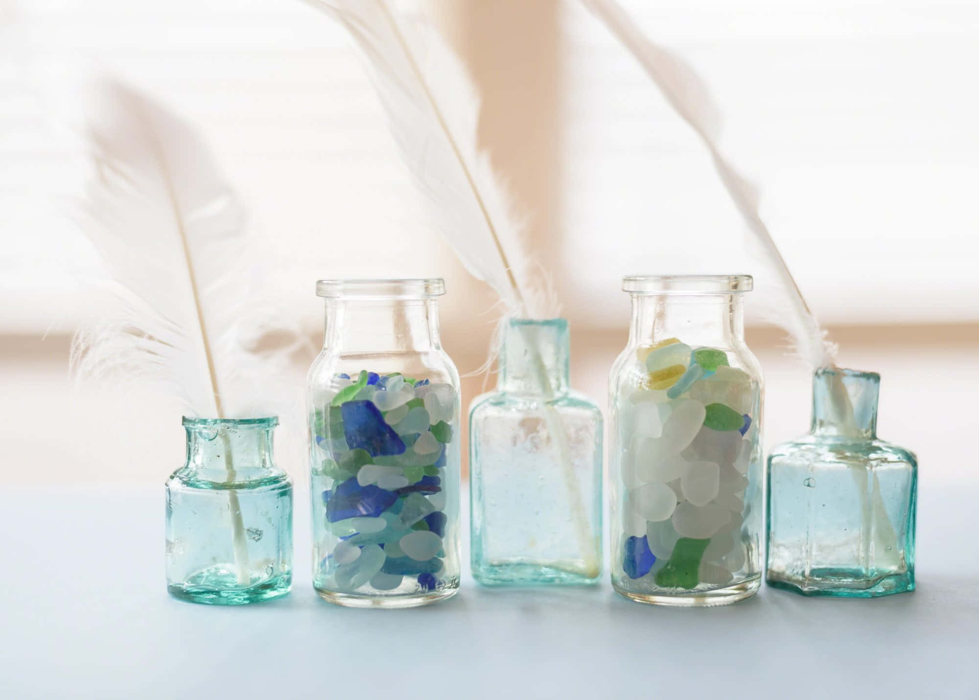 Sea Glass 桌布 壁紙 — 五個玻璃瓶裝滿彩色海玻璃和白色羽毛，在溫暖光線下放置在柔軟表面上。