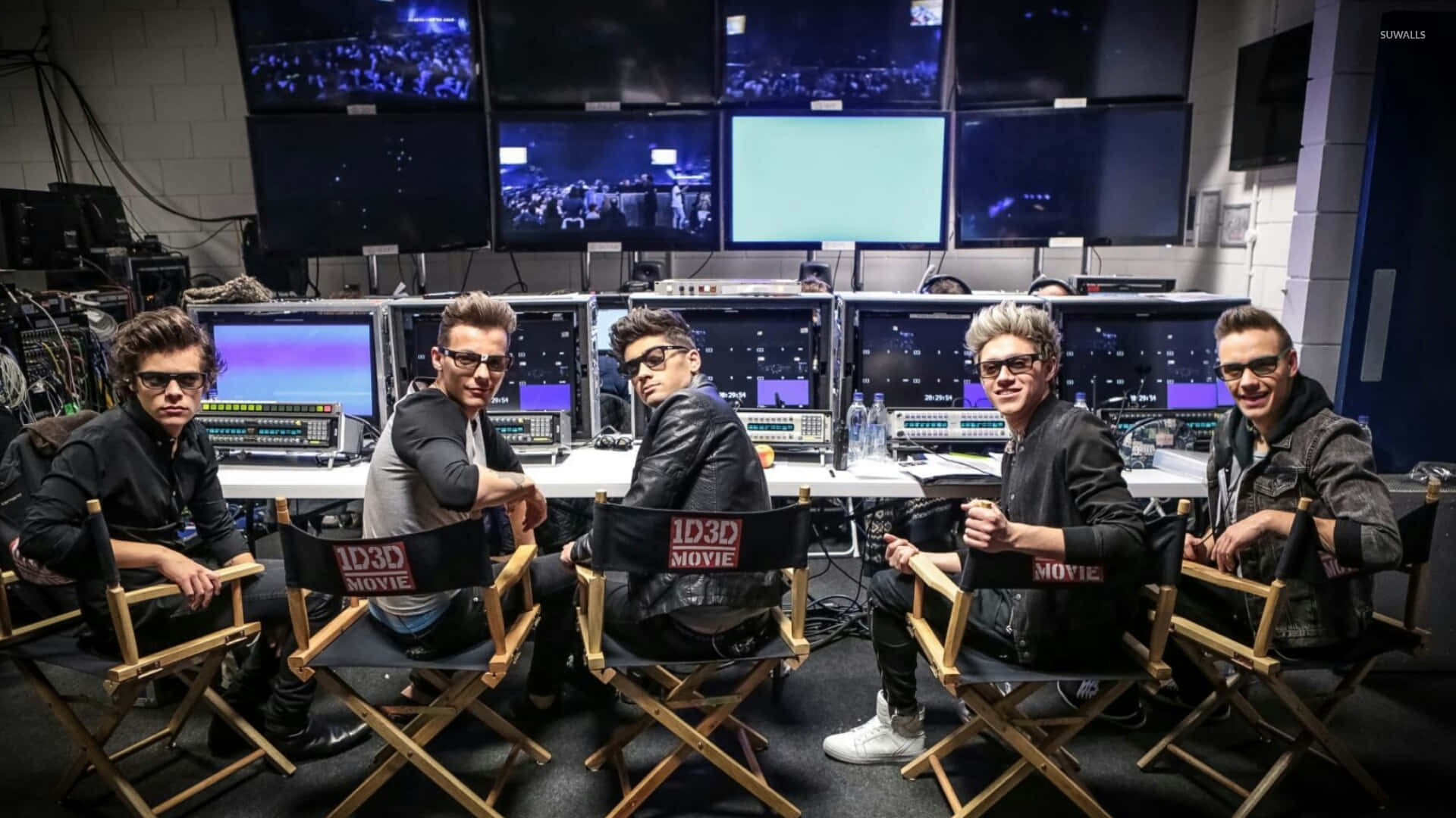 One Direction 桌布 壁紙 — 五個男人坐在導演椅上，在燈光昏暗的控制室裡自信地擺姿勢，多個顯示器播放著現場活動的畫面。