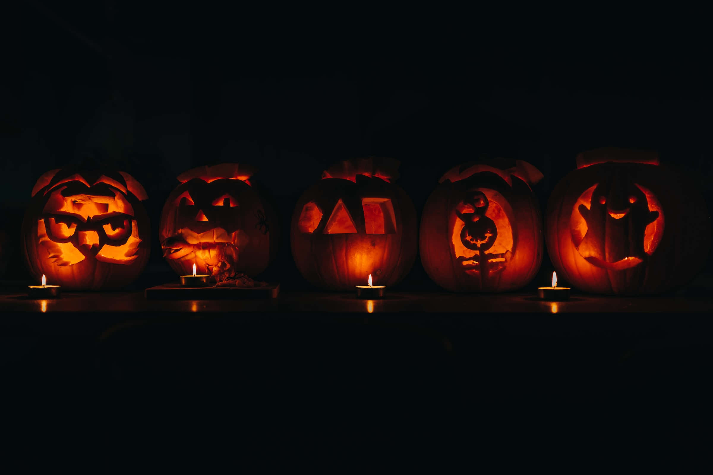 jack o'lantern 桌布 壁紙 — 五個發光的傑克南瓜燈，每個都有獨特的雕刻面孔，排成一排，在黑暗的背景中由小蠟燭照亮。