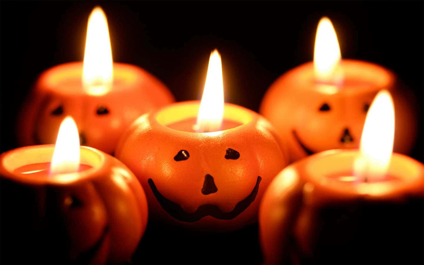 Spooky 桌布 壁紙 — 五個發光的傑克南瓜燈蠟燭，帶有歡快的面孔，在黑暗背景下溫暖地閃爍。