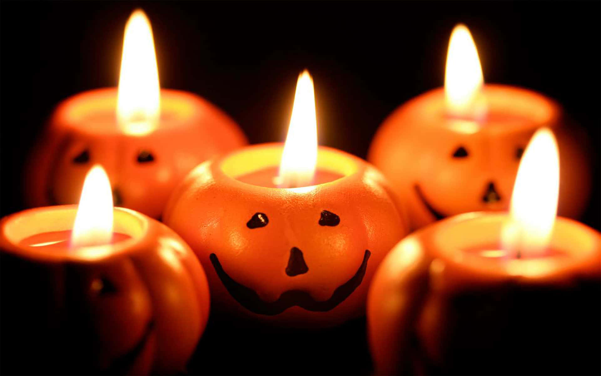 Halloween Candles 桌布 壁紙 — 五個發光的傑克南瓜燈蠟燭，帶著愉快的面孔，在黑暗背景中溫暖地閃爍。