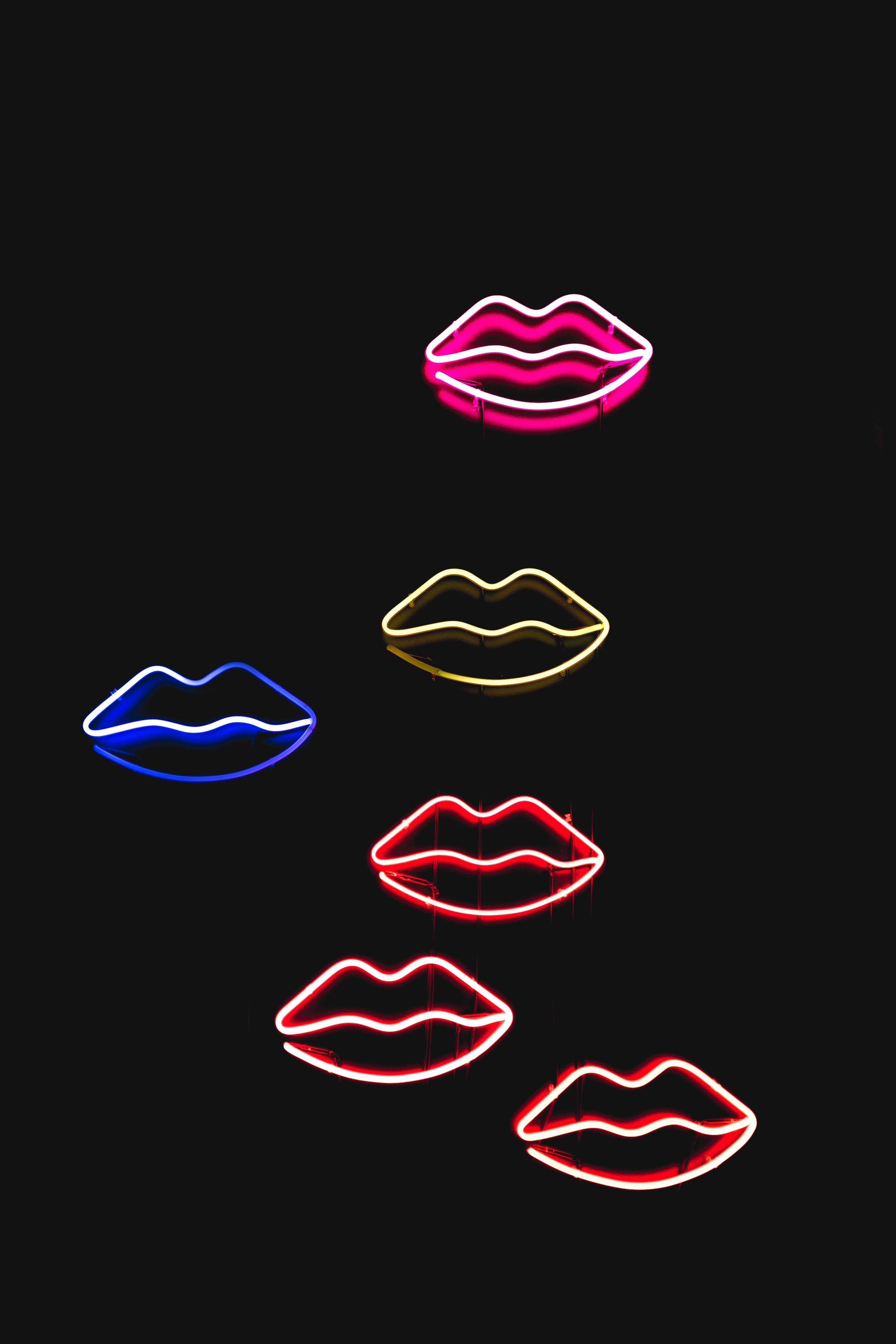 red lips 桌布 壁紙 — 五個發光的霓虹嘴唇，分別是粉紅色、藍色、黃色和紅色，漂浮在漆黑的背景上。