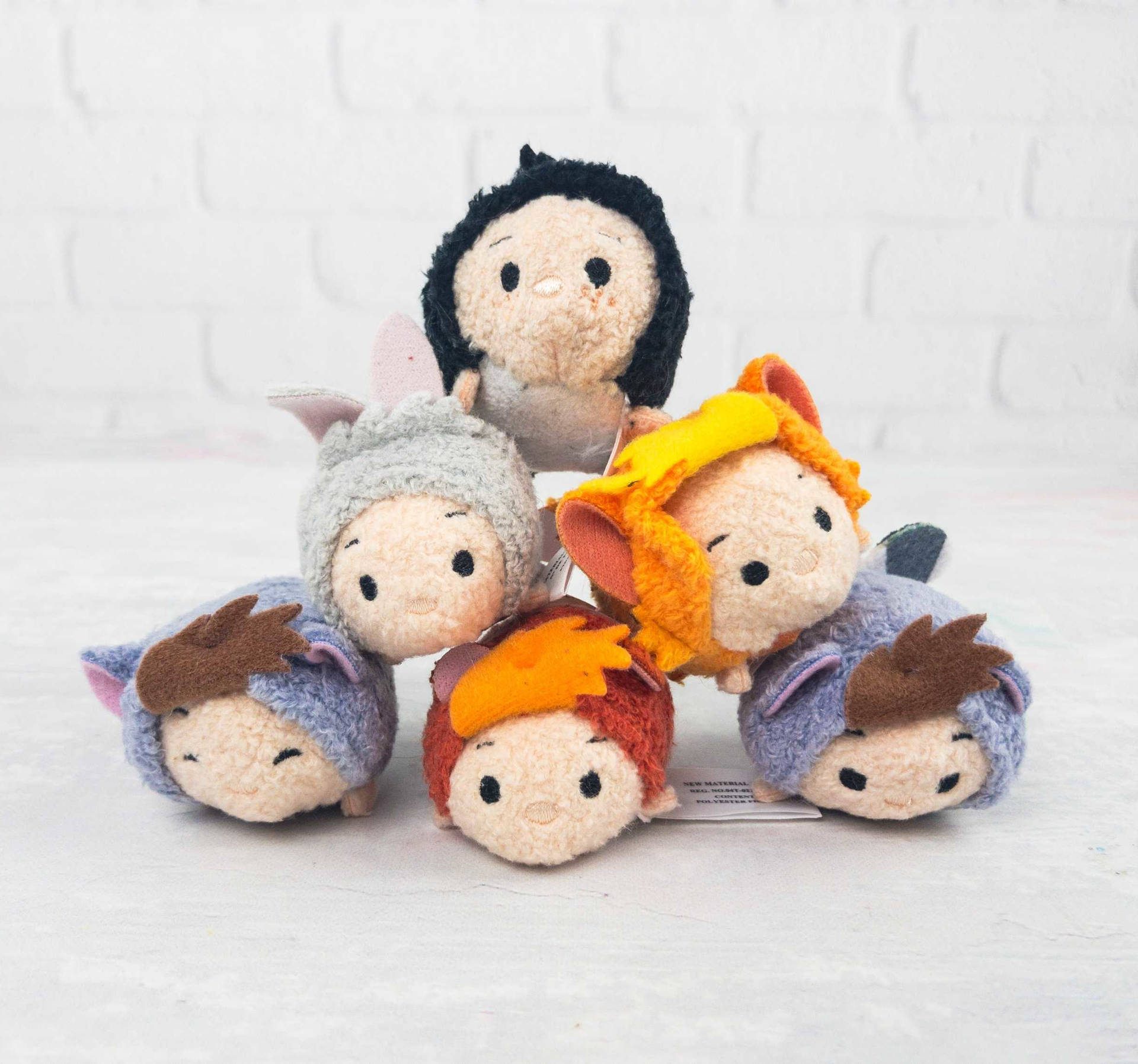 五個穿著服裝的Tsum Tsum絨毛玩具，在白色磚牆表面上排列成金字塔形狀。 — Tsum tsum 壁紙