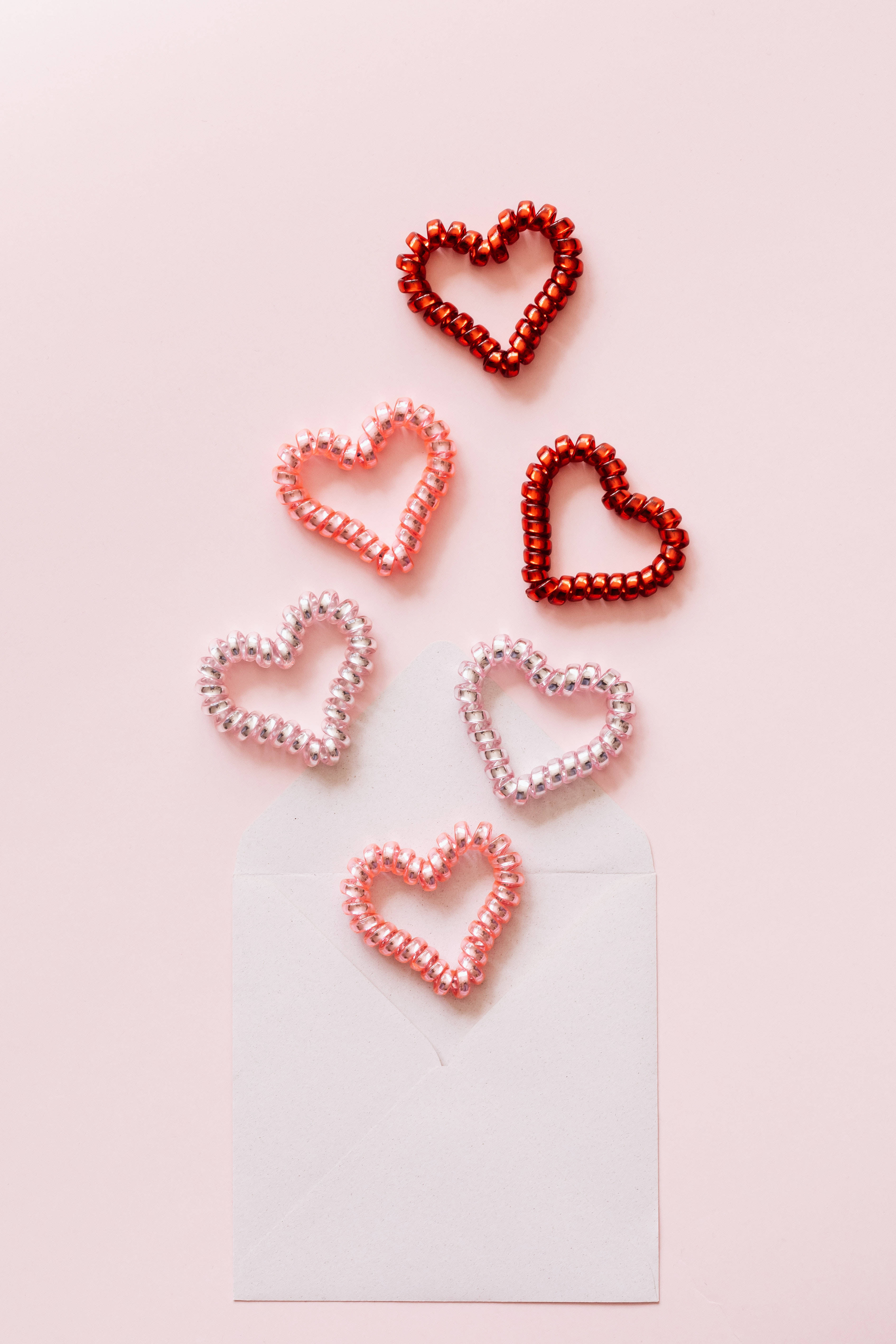Pastel Pink Heart 桌布 壁紙 — 五個紅色和粉紅色調的珠子心形漂浮在一個白色信封上方，背景是柔和的粉紅色。