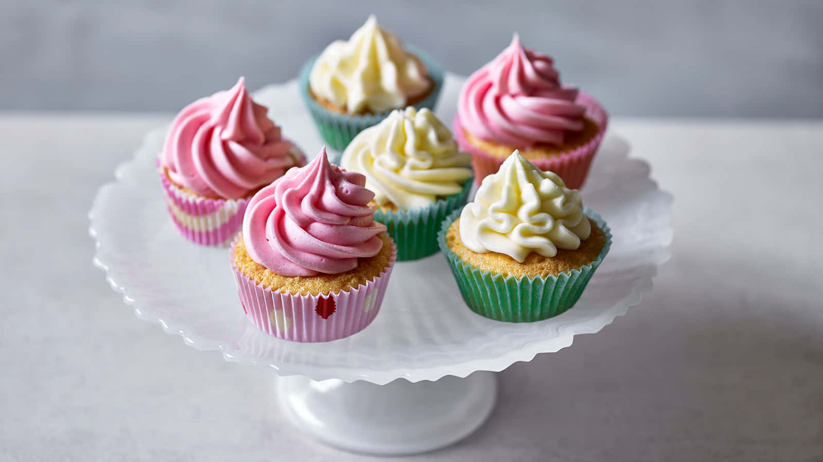 Cupcake 桌布 壁紙 — 五個色彩繽紛的杯子蛋糕，帶有粉紅色和奶油色的糖霜，優雅地放在一個白色扇貝邊蛋糕架上，背景是柔和的灰色。