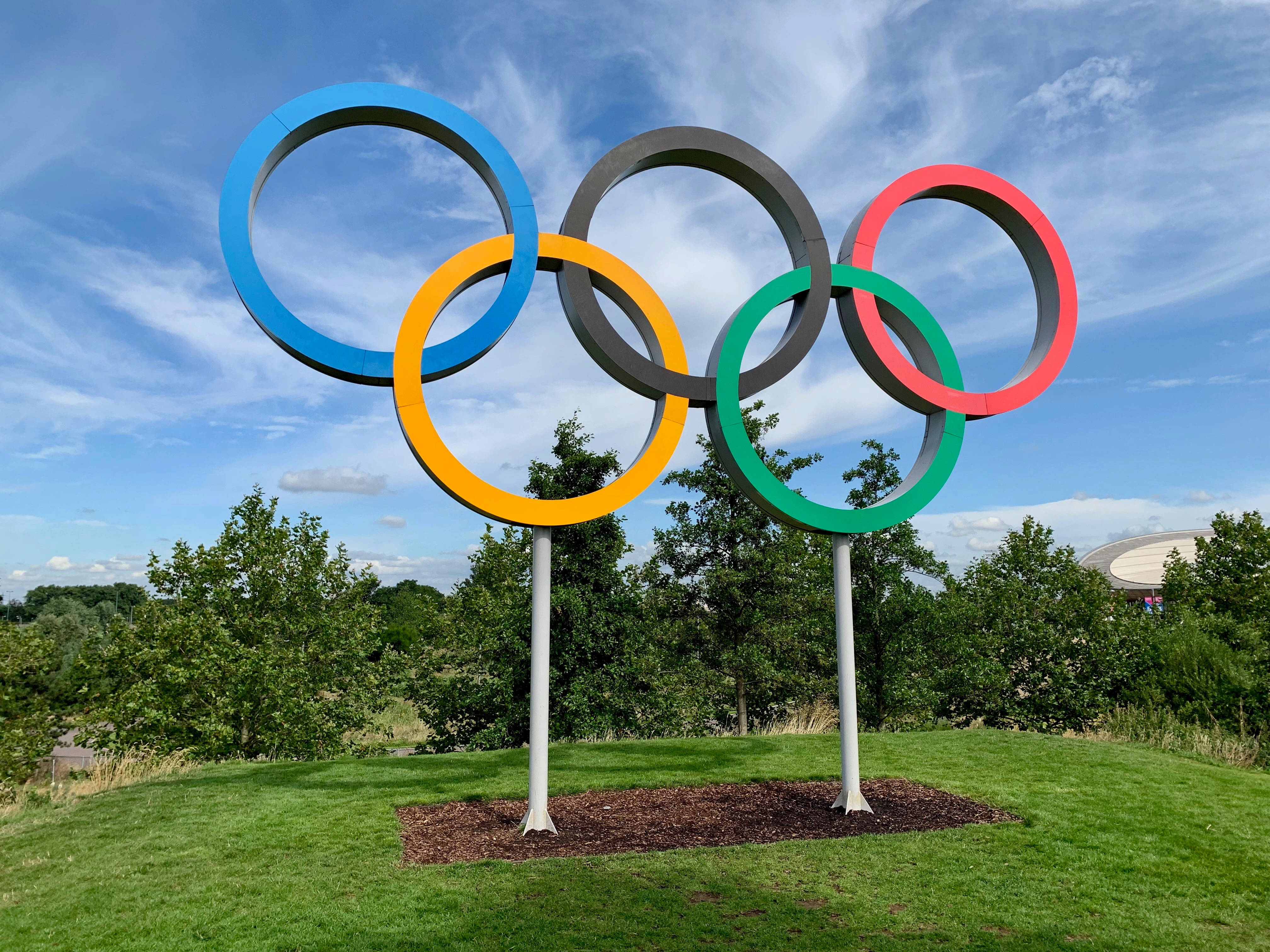 奧運 桌布 壁紙 — 五個色彩鮮豔、環環相扣的奧運五環，矗立在青草山坡上，背景是晴朗的藍天和縷縷白雲。