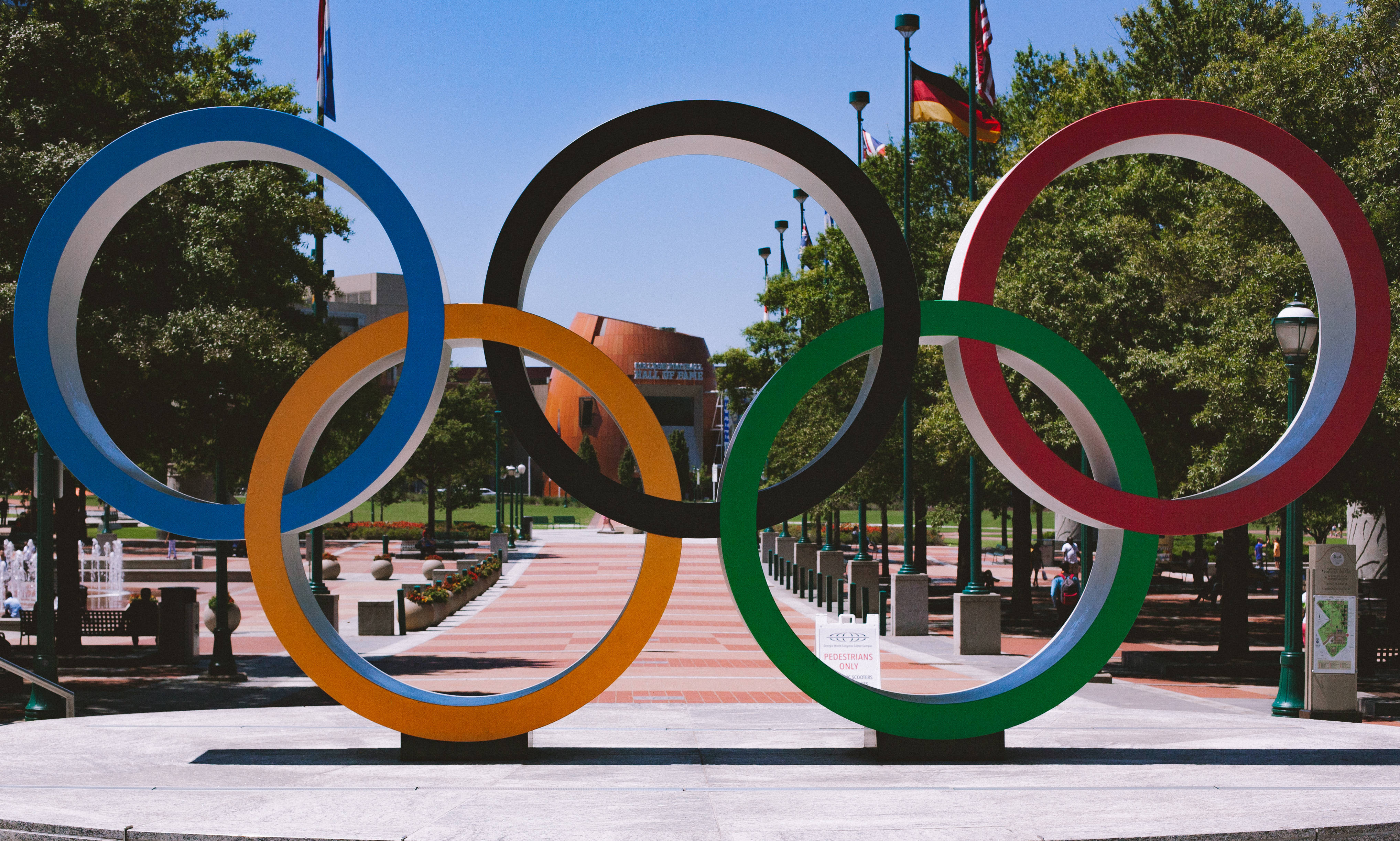 Olympics 桌布 壁紙 — 五個色彩鮮豔的奧運五環在晴朗藍天下，矗立於陽光明媚的城市公園中。