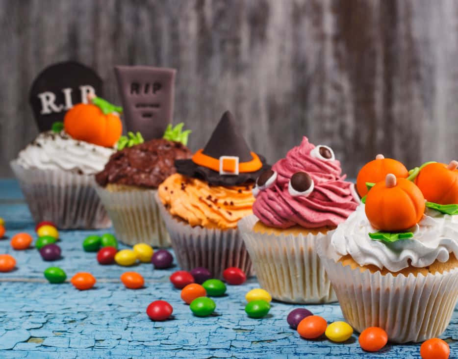 Halloween Treats 桌布 壁紙 — 五個萬聖節主題杯子蛋糕，飾有巫婆帽、南瓜和糖果眼睛，放在一個裂開的藍色木質表面上，周圍環繞著五顏六色的糖果。