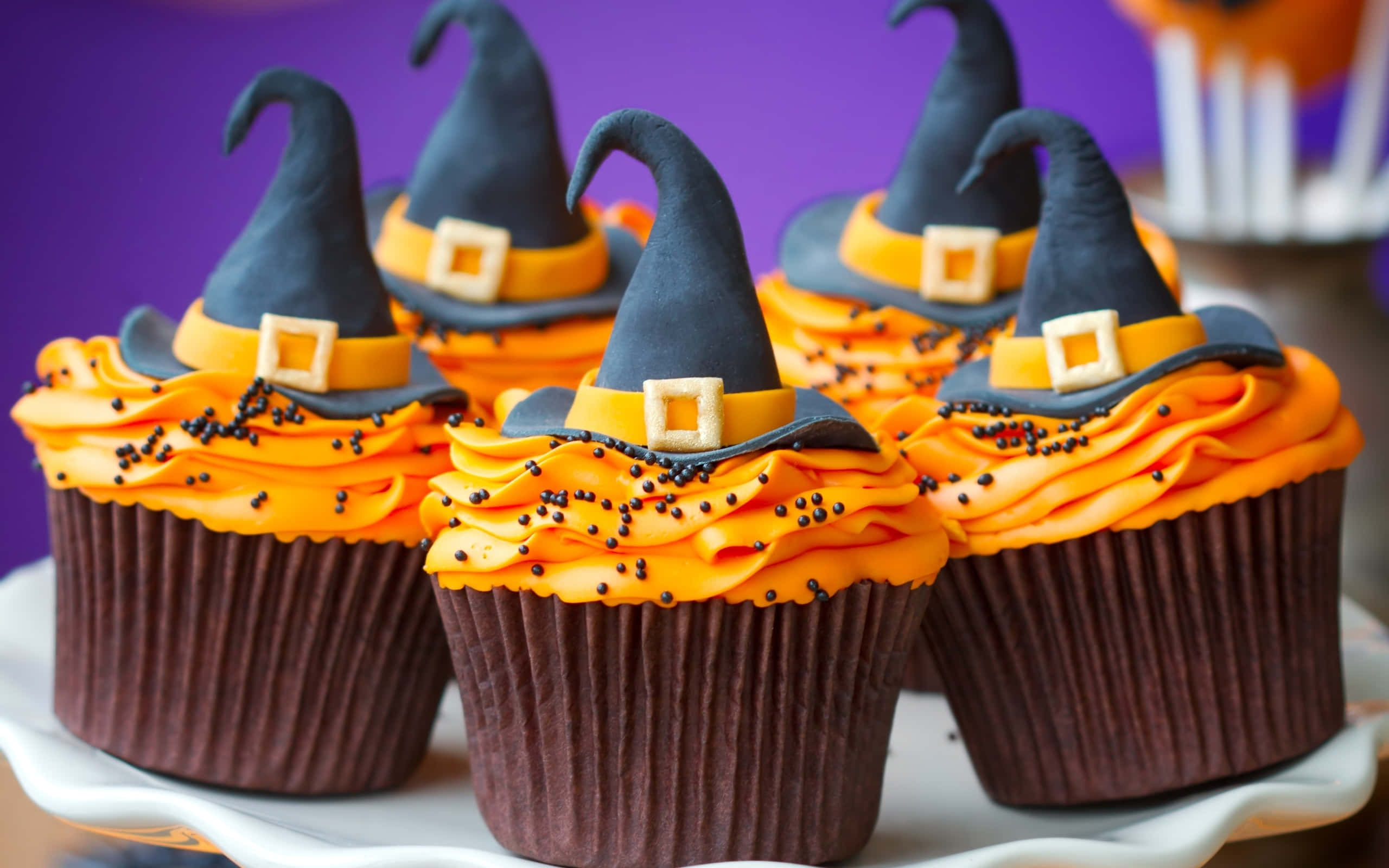 Halloween Cupcakes 桌布 壁紙 — 五個萬聖節紙杯蛋糕，頂部有巫婆帽和橙色糖霜，放在白色盤子上，背景為紫色。