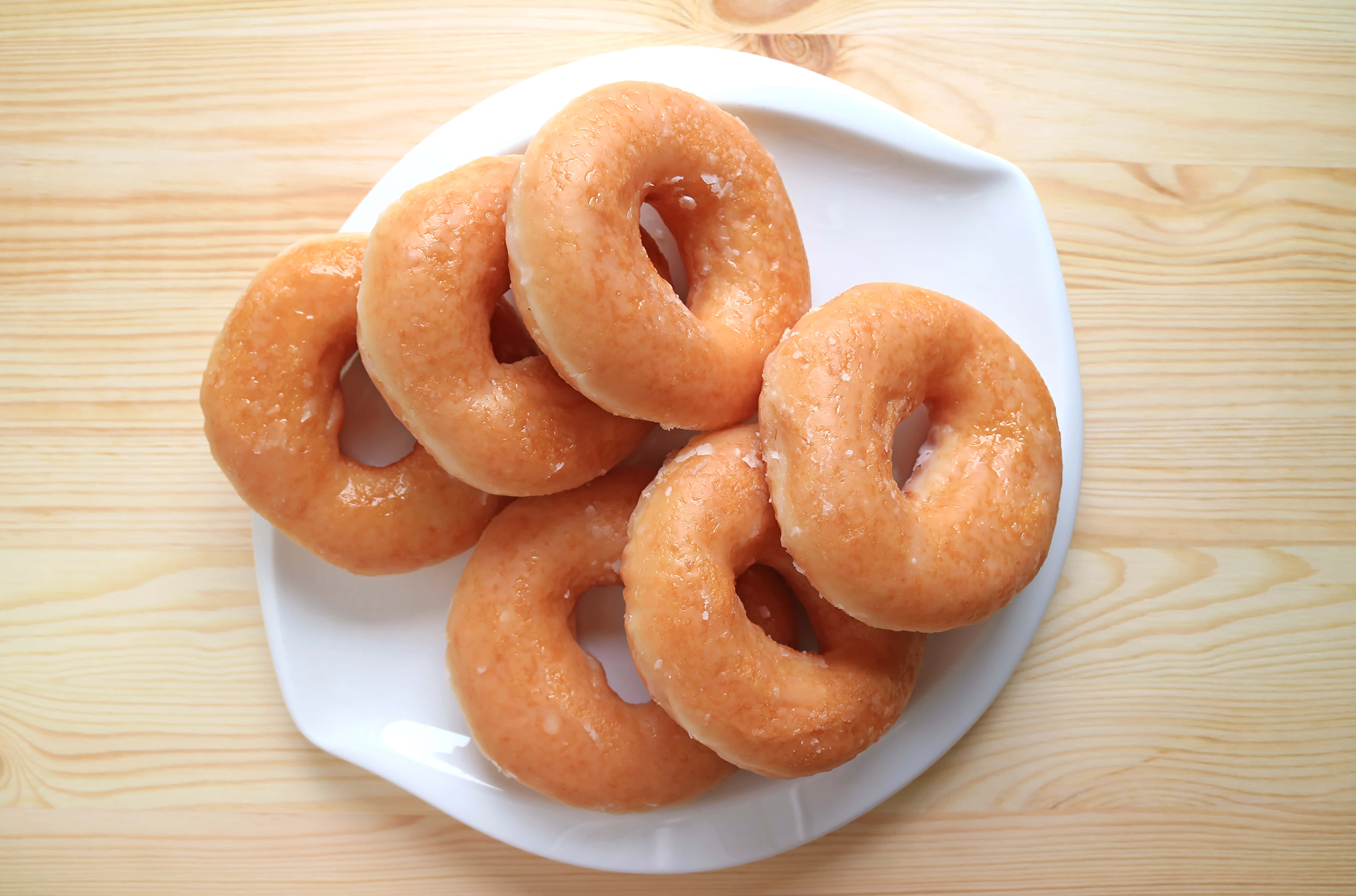 Glazed Donut 桌布 壁紙 — 五個釉麵甜甜圈堆疊在一個白色盤子上，放在淺色木質表面上。