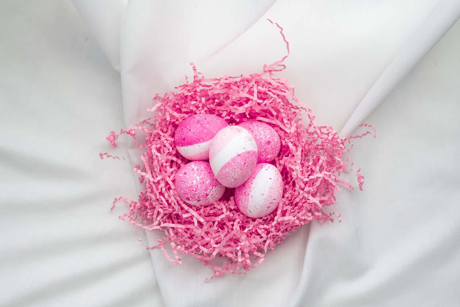 Pastel Easter 桌布 壁紙 — 五個閃閃發光的粉紅色和白色雞蛋，嵌套在充滿活力的粉紅色碎紙中，放在柔軟的白色布料上。