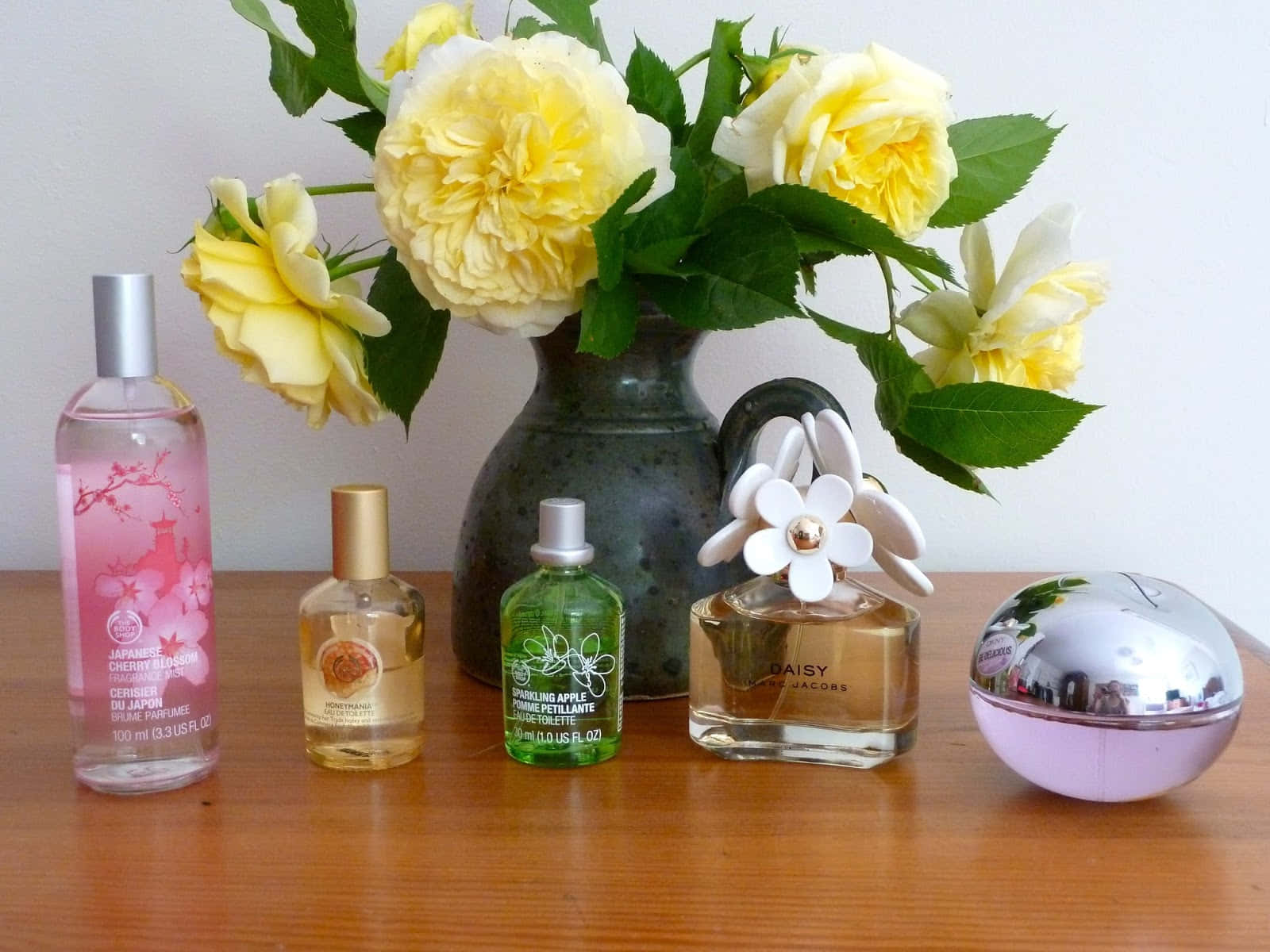 Spring Perfume 桌布 壁紙 — 五個香水瓶和一個黃玫瑰花瓶，擺放在木桌上，背景是素牆。