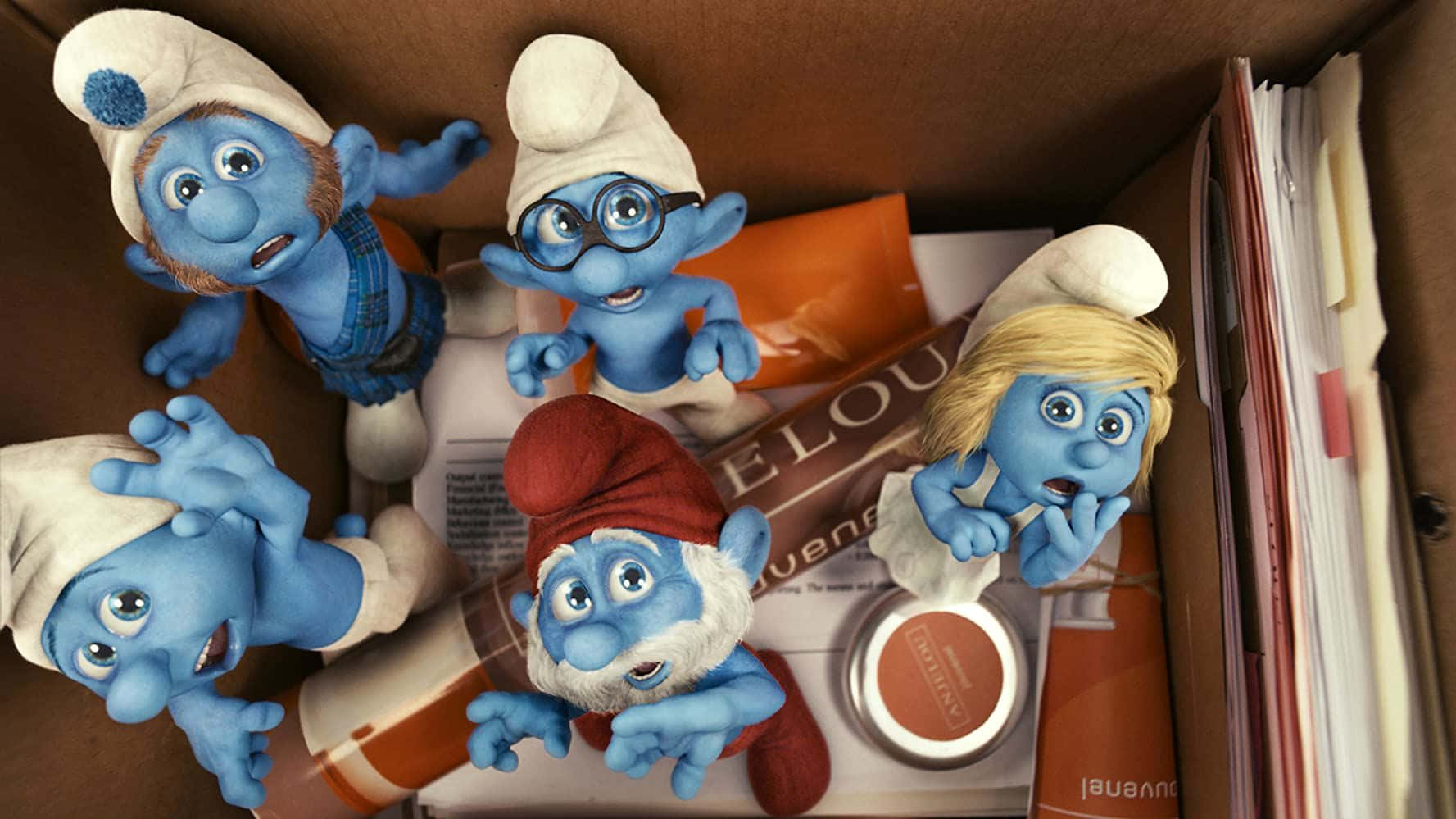 Smurf 桌布 壁紙 — 五個驚訝的藍色小精靈公仔從一個雜亂的紙箱中探出頭來，紙箱裡裝滿了紙張和管子。