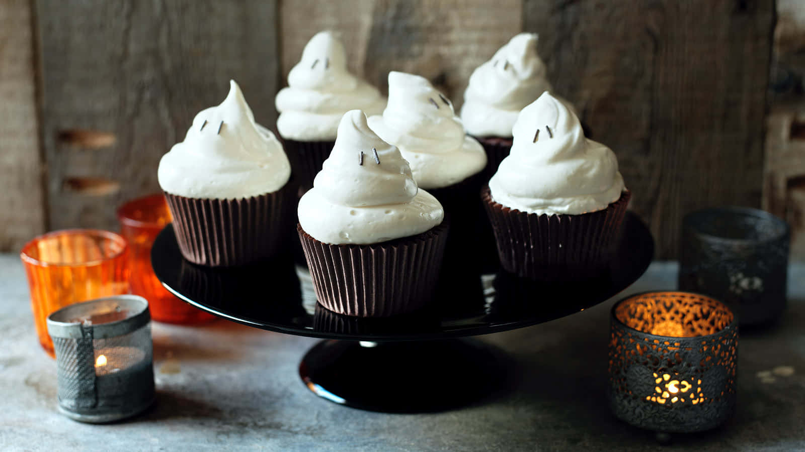 Halloween Cupcakes 桌布 壁紙 — 五個鬼臉紙杯蛋糕，頂部有白色蛋白霜，放在一個黑色蛋糕架上，周圍環繞著發光的蠟燭。