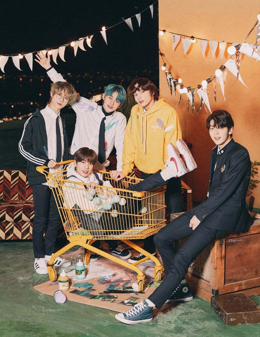 Big Hit Entertainment 桌布 壁紙 — 五名年輕男子在夜晚的串燈下圍著一架黃色購物車擺出俏皮的姿勢，周圍散落著繪畫用品。