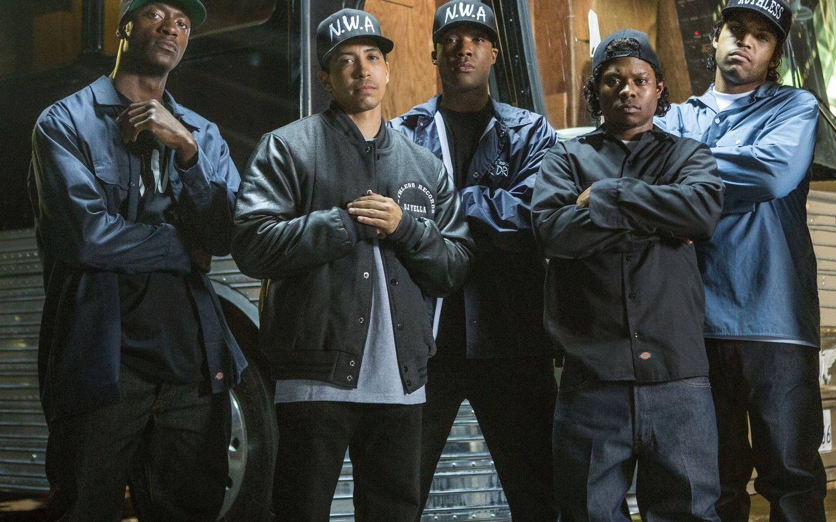 Ice Cube 桌布 壁紙 — 五名戴著N.W.A帽子和穿著外套的男子在昏暗的車庫燈光下自信地站在一輛老爺車前。