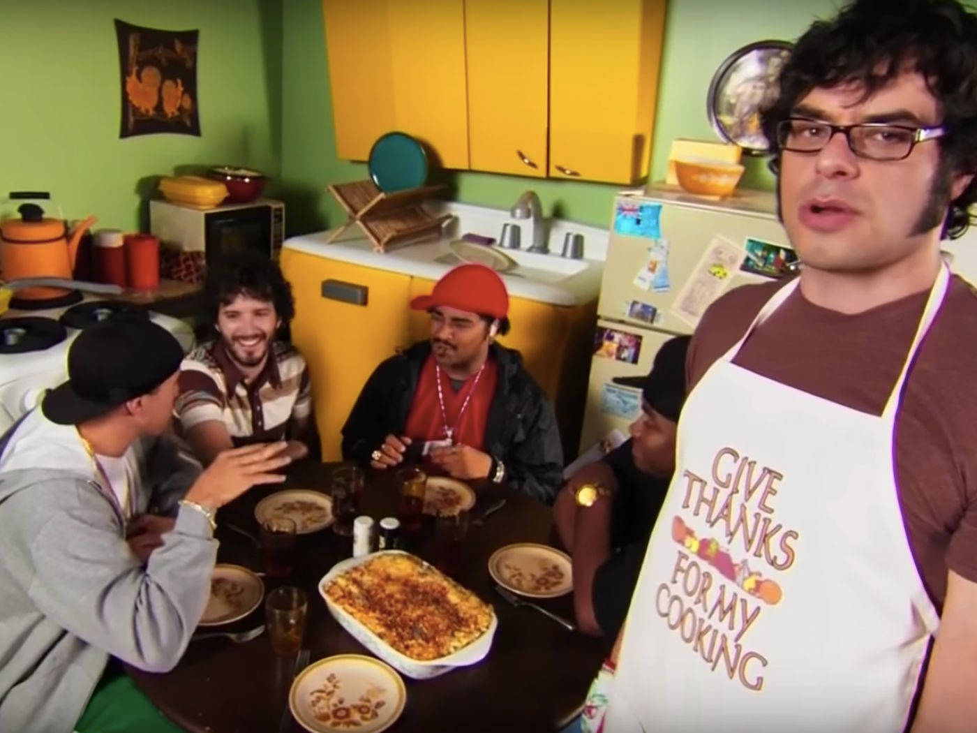 Flight Of The Conchords 桌布 壁紙 — 五名男子圍在廚房餐桌旁享用美食，其中一名穿著圍裙的男子自豪地展示「Give Thanks For My Cooking」。