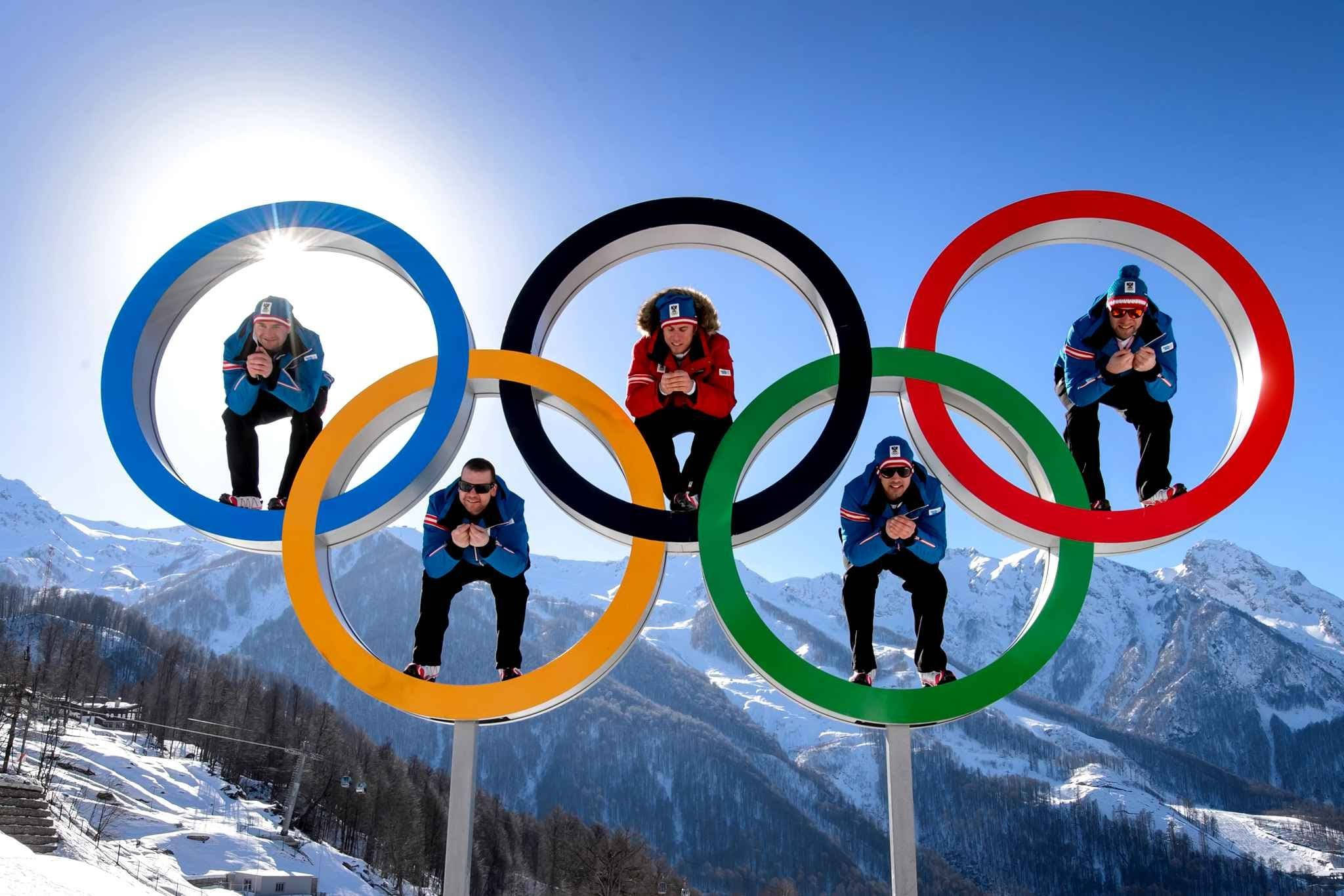 Winter Olympics 桌布 壁紙 — 五名穿著冬季裝備的運動員在奧運五環內擺姿勢，背景是明亮的藍天和白雪覆蓋的山脈。