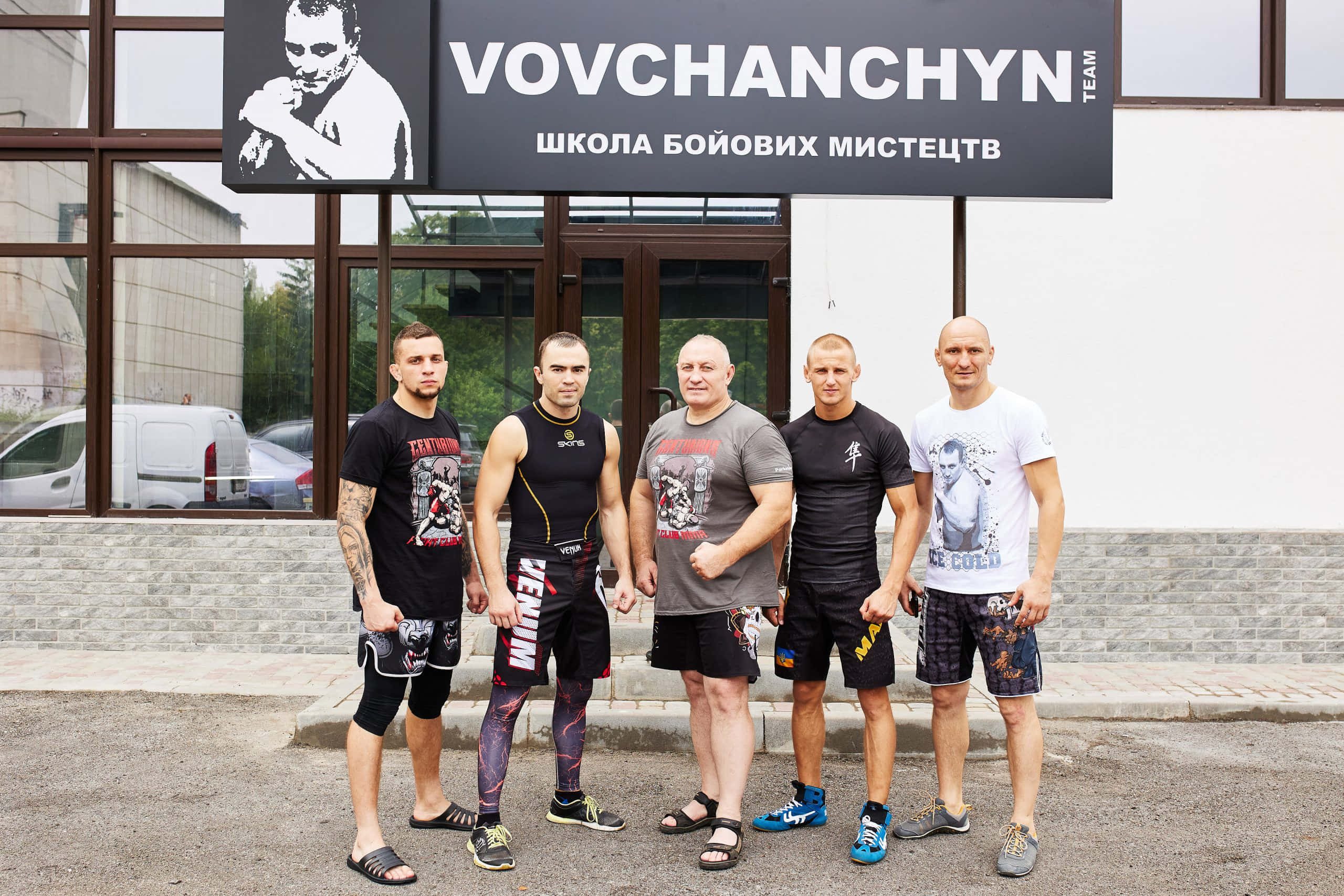 Fight 桌布 壁紙 — 五名肌肉發達的男子身穿戰鬥裝備，自信地站在一間武術館外，旁邊有醒目的Vovchanchyn團隊標誌。