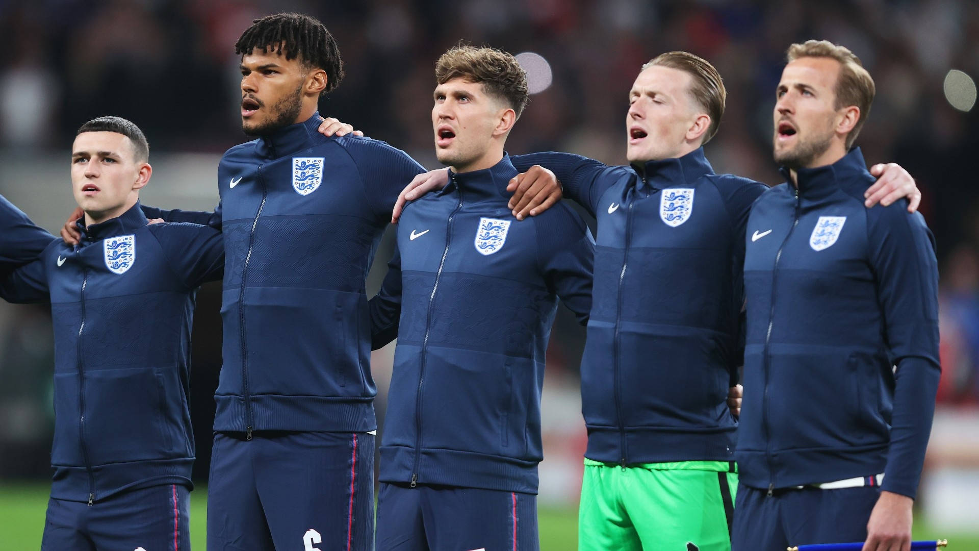 England 桌布 壁紙 — 五名身穿海軍藍運動服的英格蘭足球運動員在球場上肩並肩站著，張開嘴巴，手臂相連，唱著國歌。