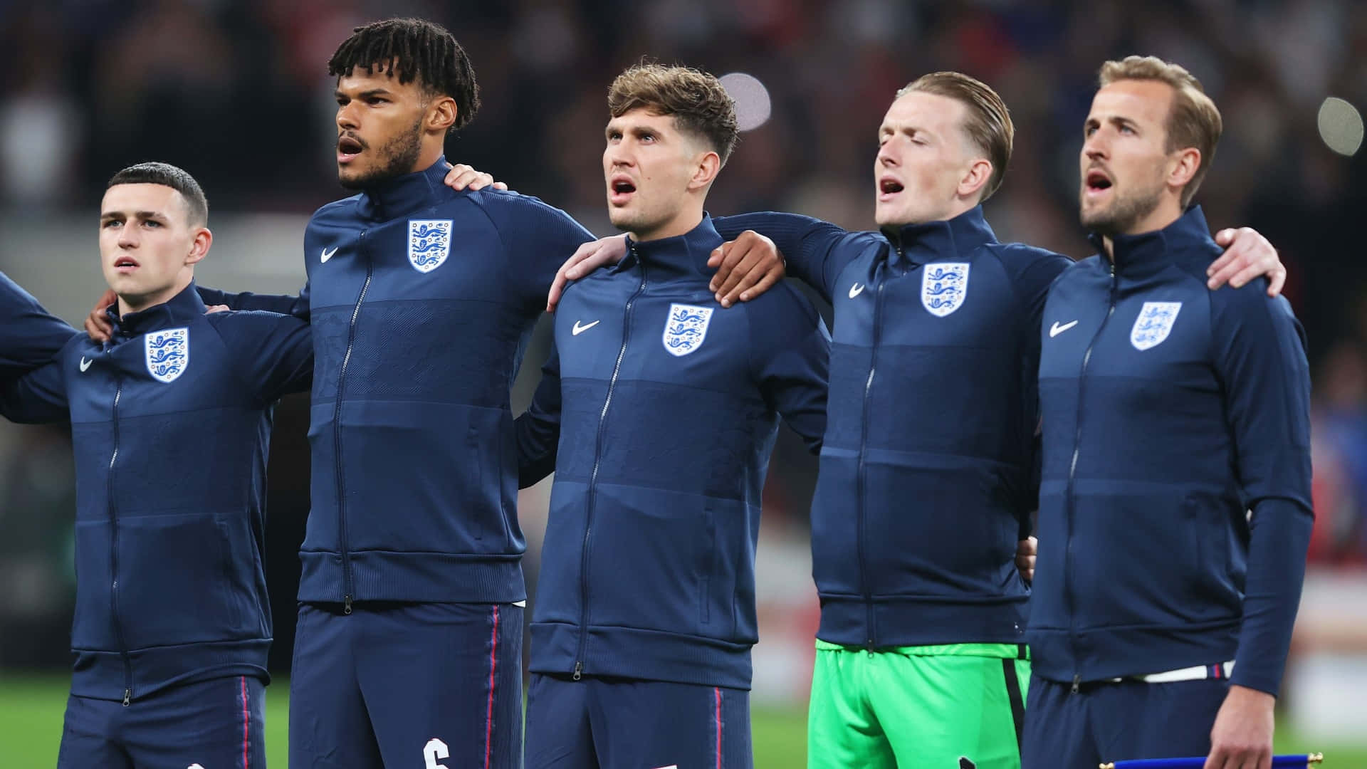 Football Players 桌布 壁紙 — 五名身穿海軍藍運動服的英格蘭足球員在球場上肩並肩站立，張開嘴巴，手臂相連，高唱國歌。
