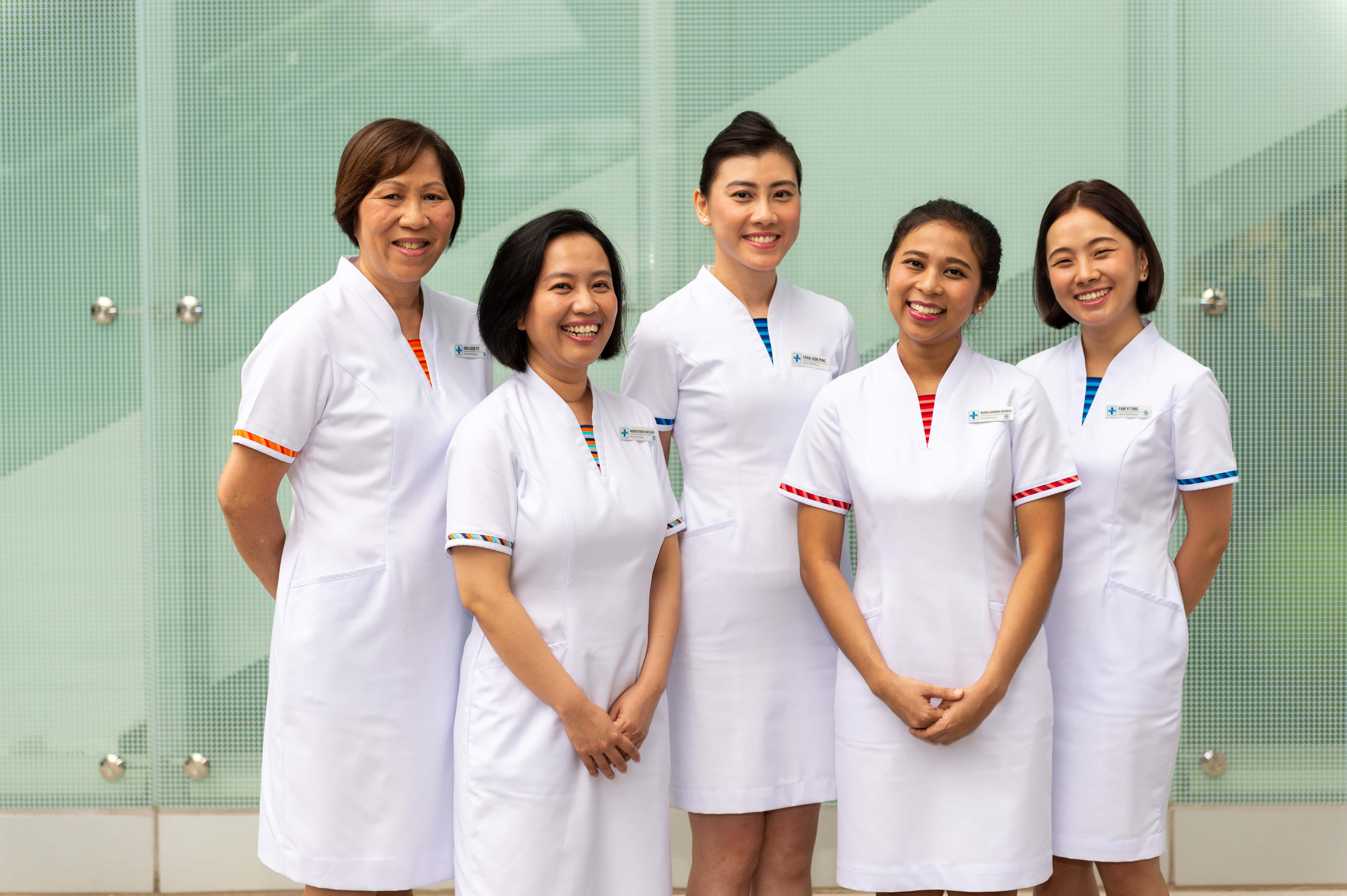 Nursing 桌布 壁紙 — 五名身穿白色制服、面帶微笑的醫護人員站在現代玻璃牆前。
