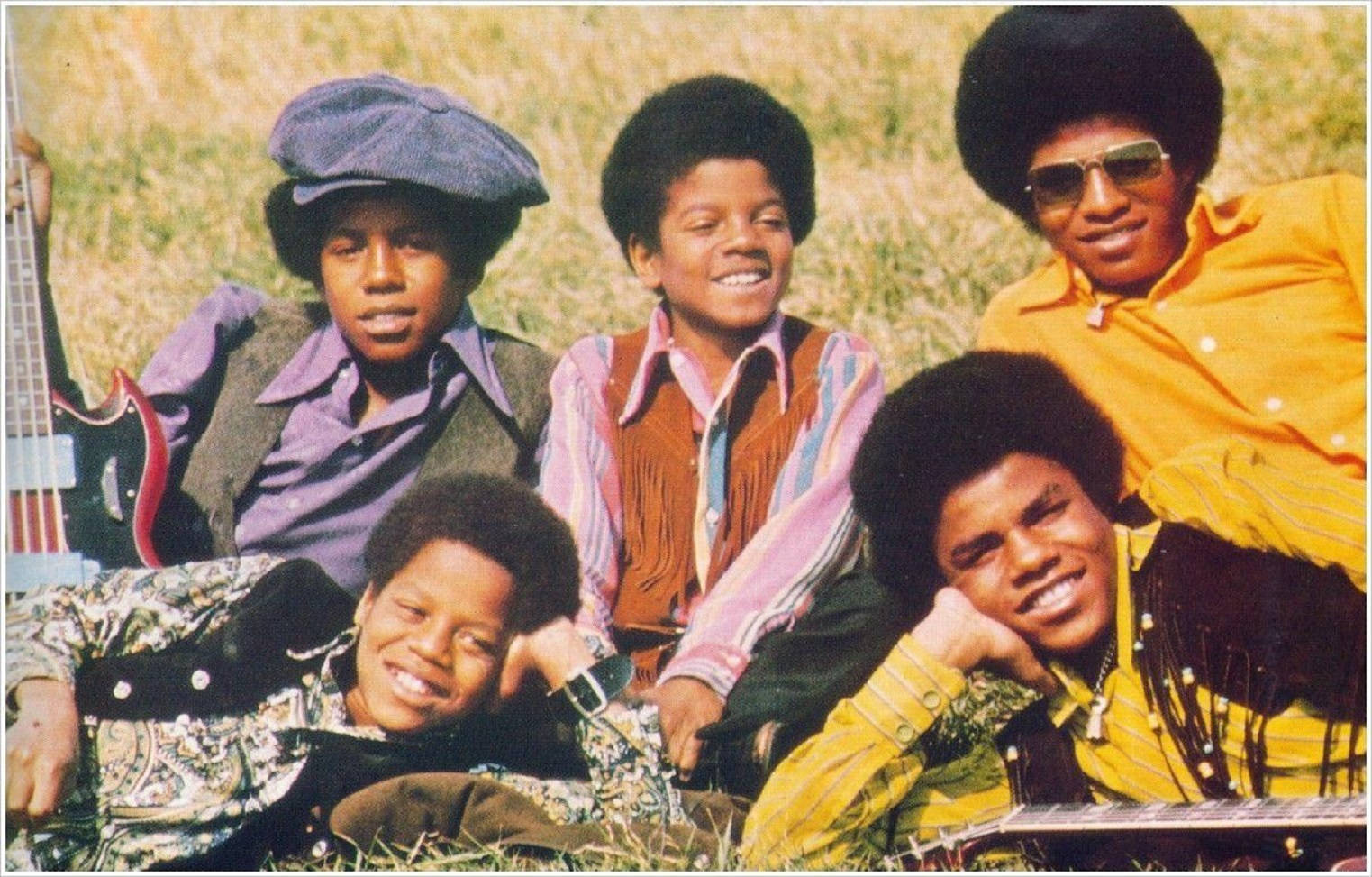 麥可·傑克森 桌布 壁紙 — 五名身穿鮮豔1970年代服裝的年輕男子在陽光明媚的草地上微笑並隨意擺姿勢，手持吉他。