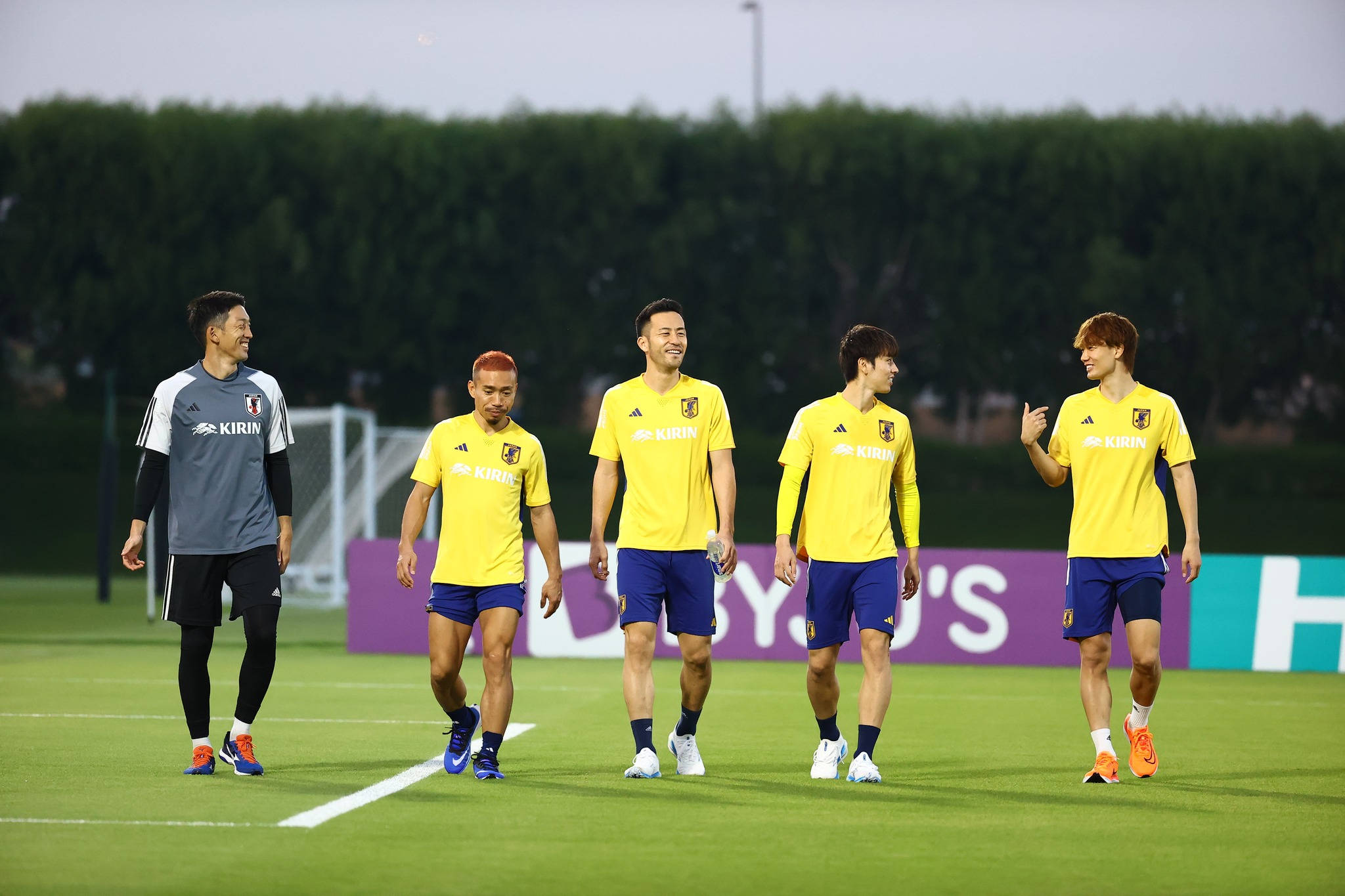 Football Players 桌布 壁紙 — 五名身穿黃色球衣的日本足球員在訓練期間走過綠色球場，一邊聊天一邊微笑。