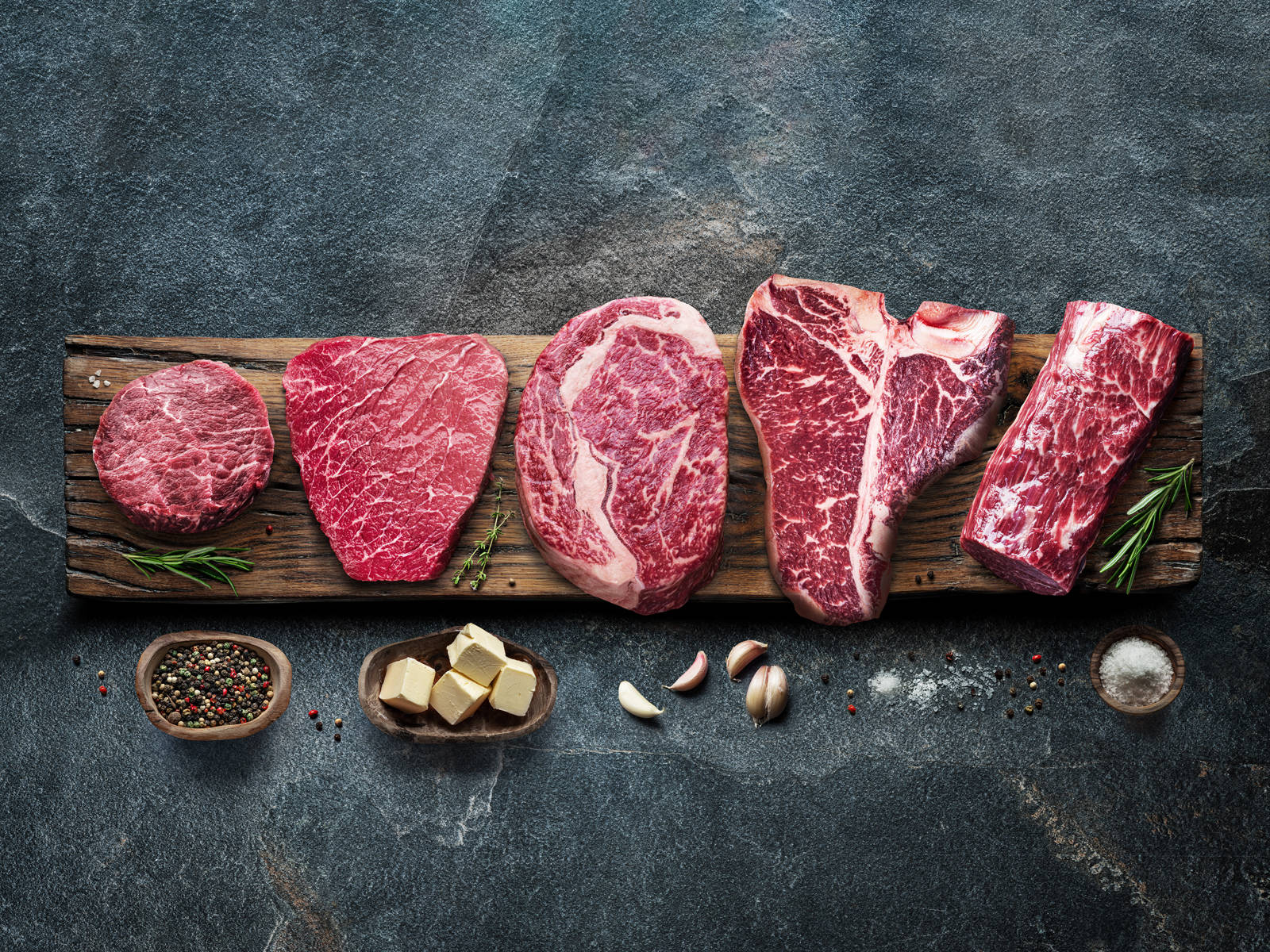 meat 桌布 壁紙 — 五塊優質生牛肉排列在質樸的木板上，搭配香草、奶油、大蒜和調味料，背景為深色石頭。