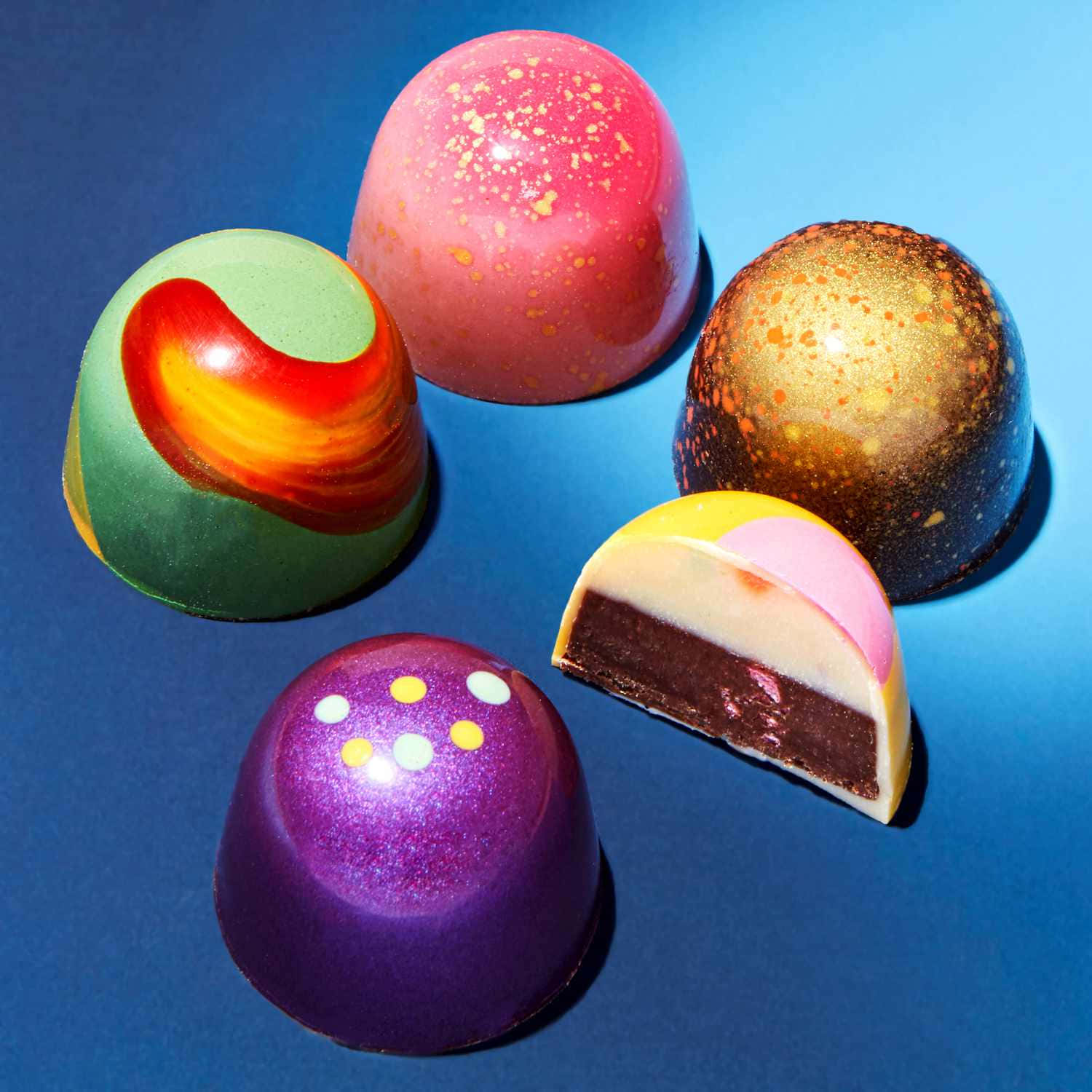 Japanese Sweets 桌布 壁紙 — 五塊光滑、色彩鮮豔的圓頂形巧克力，帶有亮粉和漩渦圖案，放在深藍色背景上，其中一塊被切開，露出層次分明的內部。