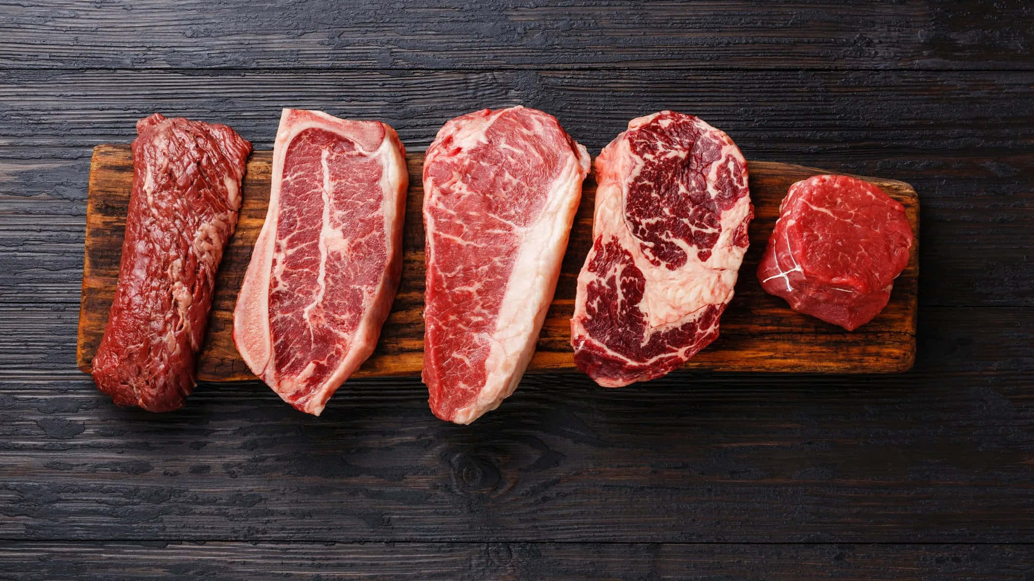 meat 桌布 壁紙 — 五塊帶有大理石紋的生牛肉，排列在木板上，背景為深色鄉村風格木材。