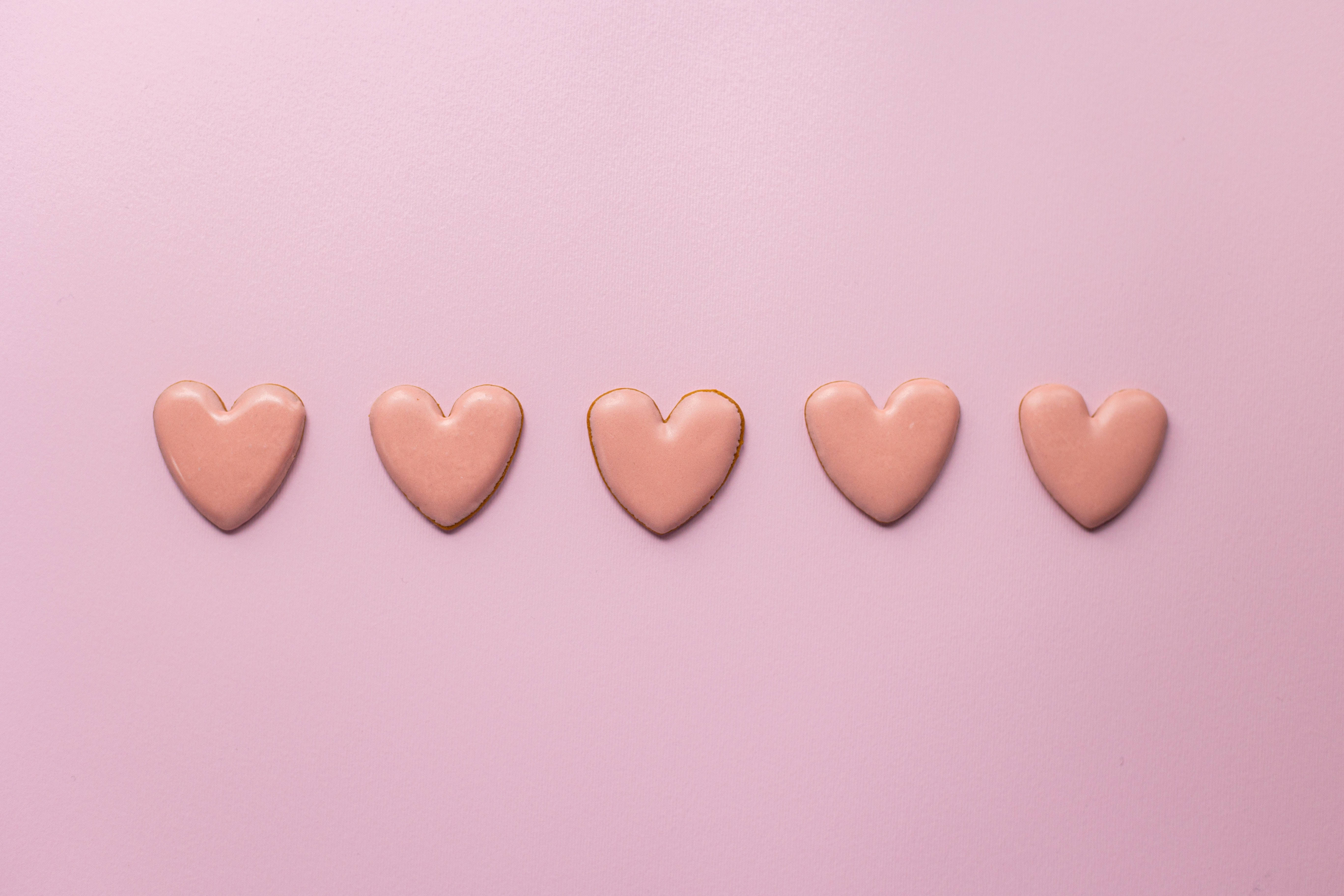 Pastel Pink Heart 桌布 壁紙 — 五塊心形餅乾，上面有粉紅色糖霜，整齊地排列在柔和的粉紅色背景上。