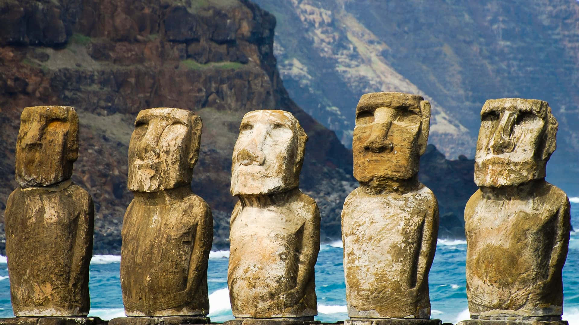 Chile 桌布 壁紙 — 五尊古老的石像摩艾石像莊嚴地矗立在懸崖邊，俯瞰著海洋，背後是崎嶇的山脈。