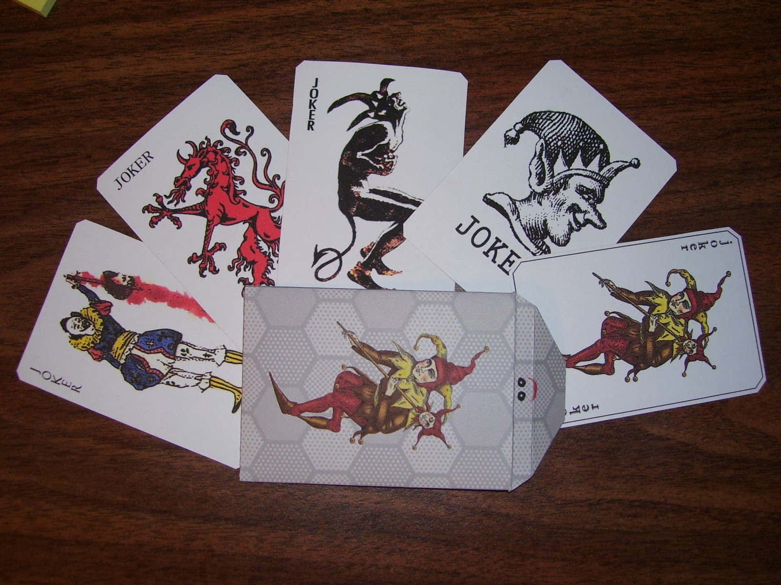 Joker Card 桌布 壁紙 — 五張插圖小丑撲克牌在深色木桌上扇形展開，展現奇思妙想與惡魔般的設計。