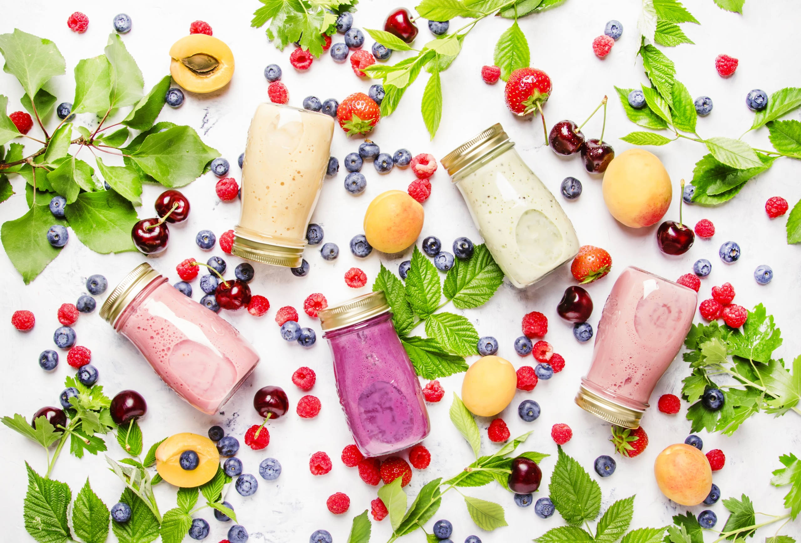 Smoothie 桌布 壁紙 — 五杯色彩繽紛的冰沙放在玻璃罐中，周圍是新鮮漿果和綠葉，置於白色表面上。