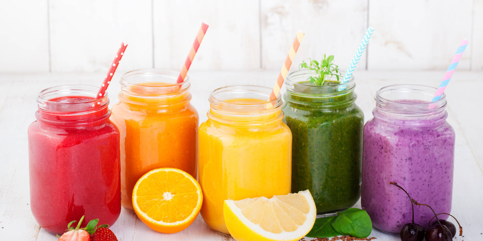 Smoothie 桌布 壁紙 — 五杯色彩鮮豔的冰沙放在梅森罐中，配有條紋吸管，周圍是新鮮水果，置於白色木質表面上。