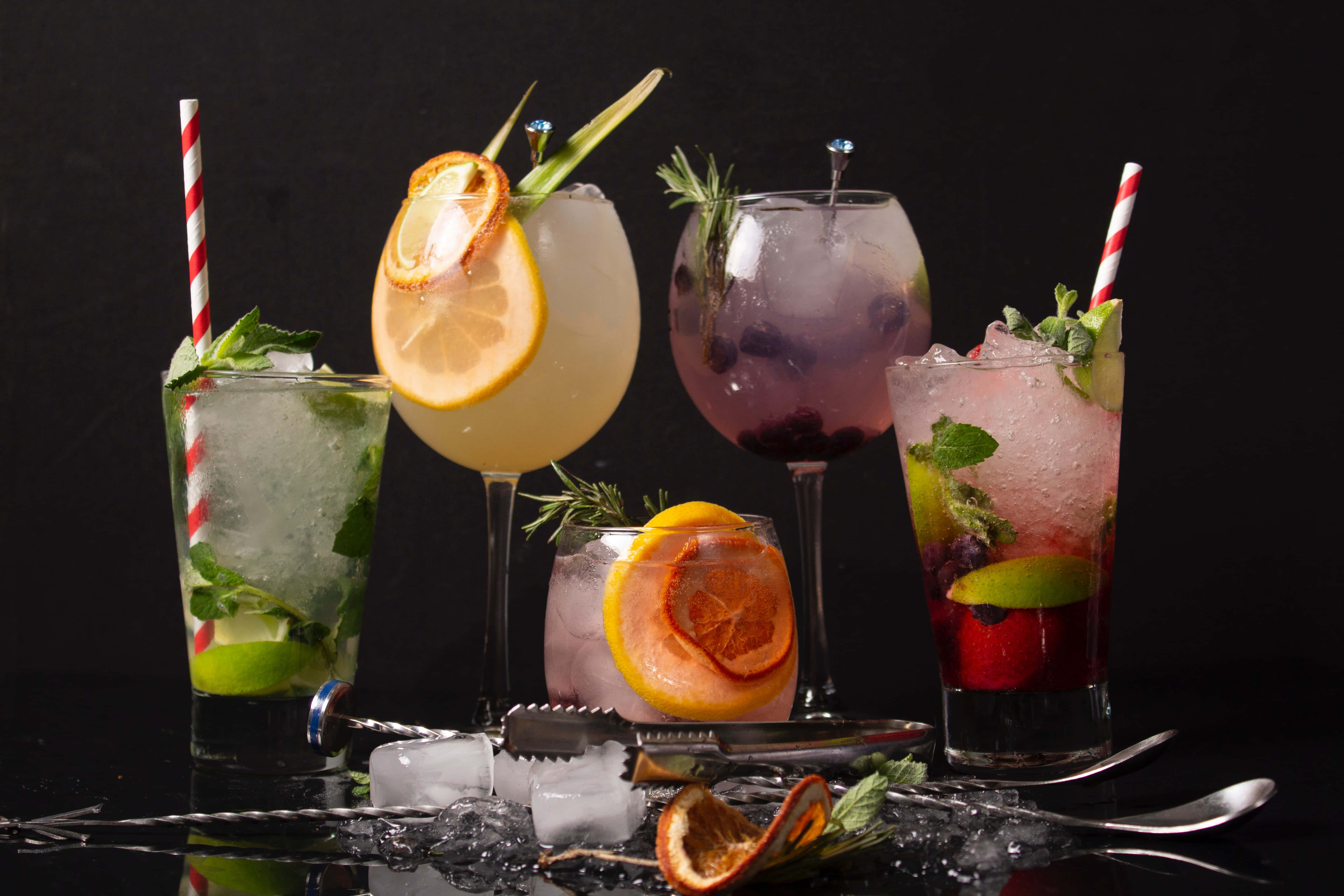 Summer Drinks 桌布 壁紙 — 五杯色彩鮮豔的雞尾酒，配上柑橘裝飾和薄荷，放在黑色表面上，周圍有冰塊和酒吧工具。