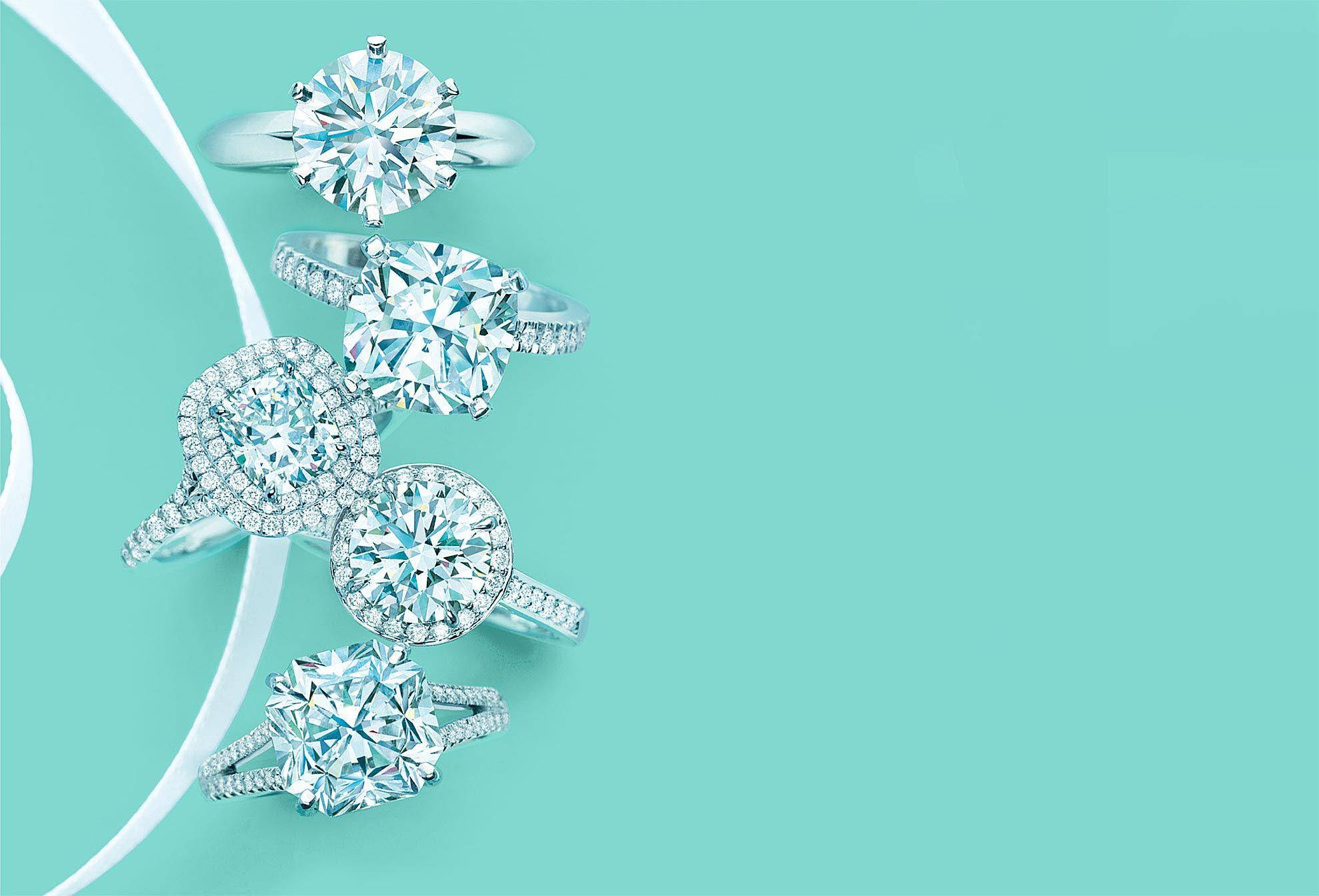 tiffany 桌布 壁紙 — 五枚閃爍的鑽石戒指搭配銀色戒環，在充滿活力的藍綠色背景上以對角線排列，並帶有白色絲帶曲線。