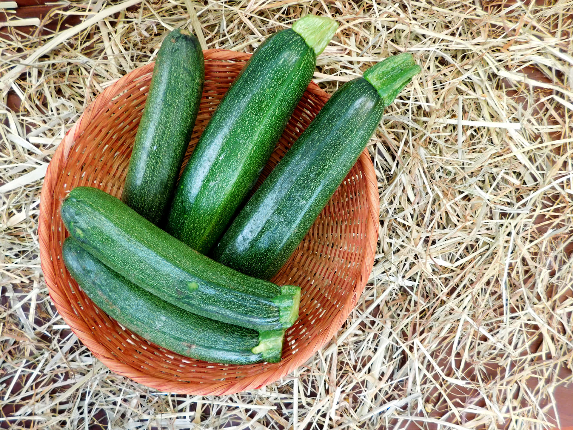 Zuchinis 壁紙 — 五條光滑的綠色櫛瓜放在一個編織的橙色籃子裡，籃子置於乾草堆上。