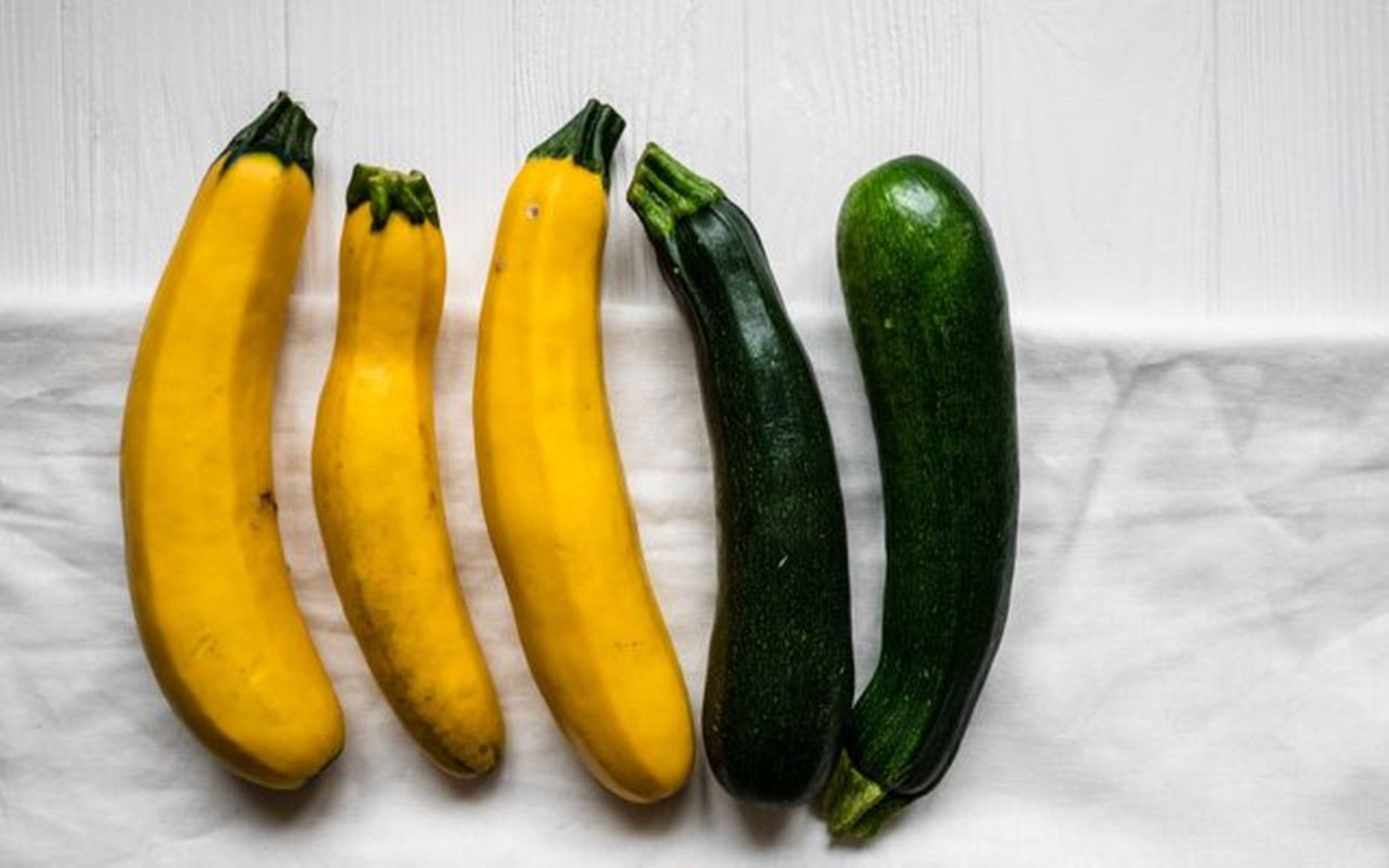 Vegetables 桌布 壁紙 — 五條新鮮的黃色和綠色櫛瓜整齊地排列在白色布上，背景是淺色木紋。