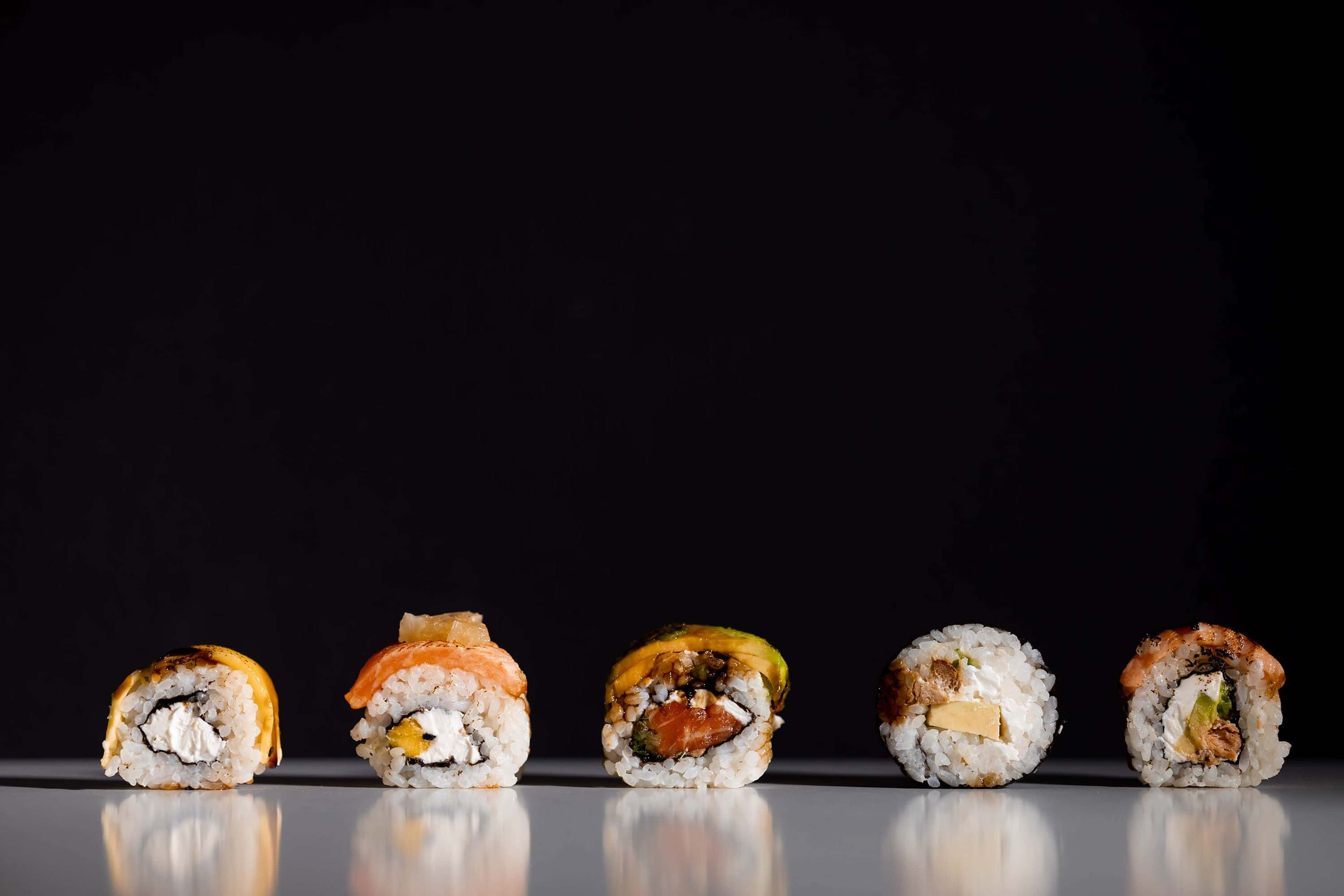Sushi 桌布 壁紙 — 五款色彩繽紛的壽司卷排列成一排，放在黑色背景前的反光表面上。